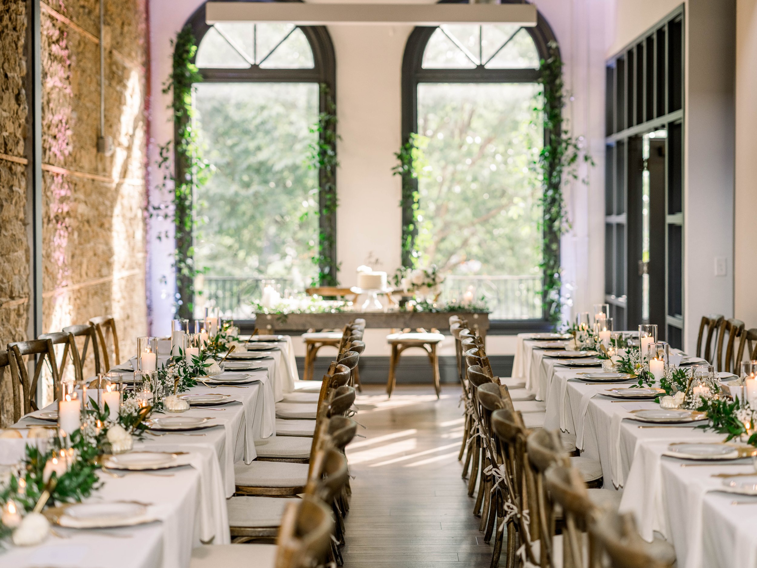 Wedding reception setup with long tables decorated with white tablecloths, candles, and greenery, in a room with large arched windows and a stone wall on the left side.