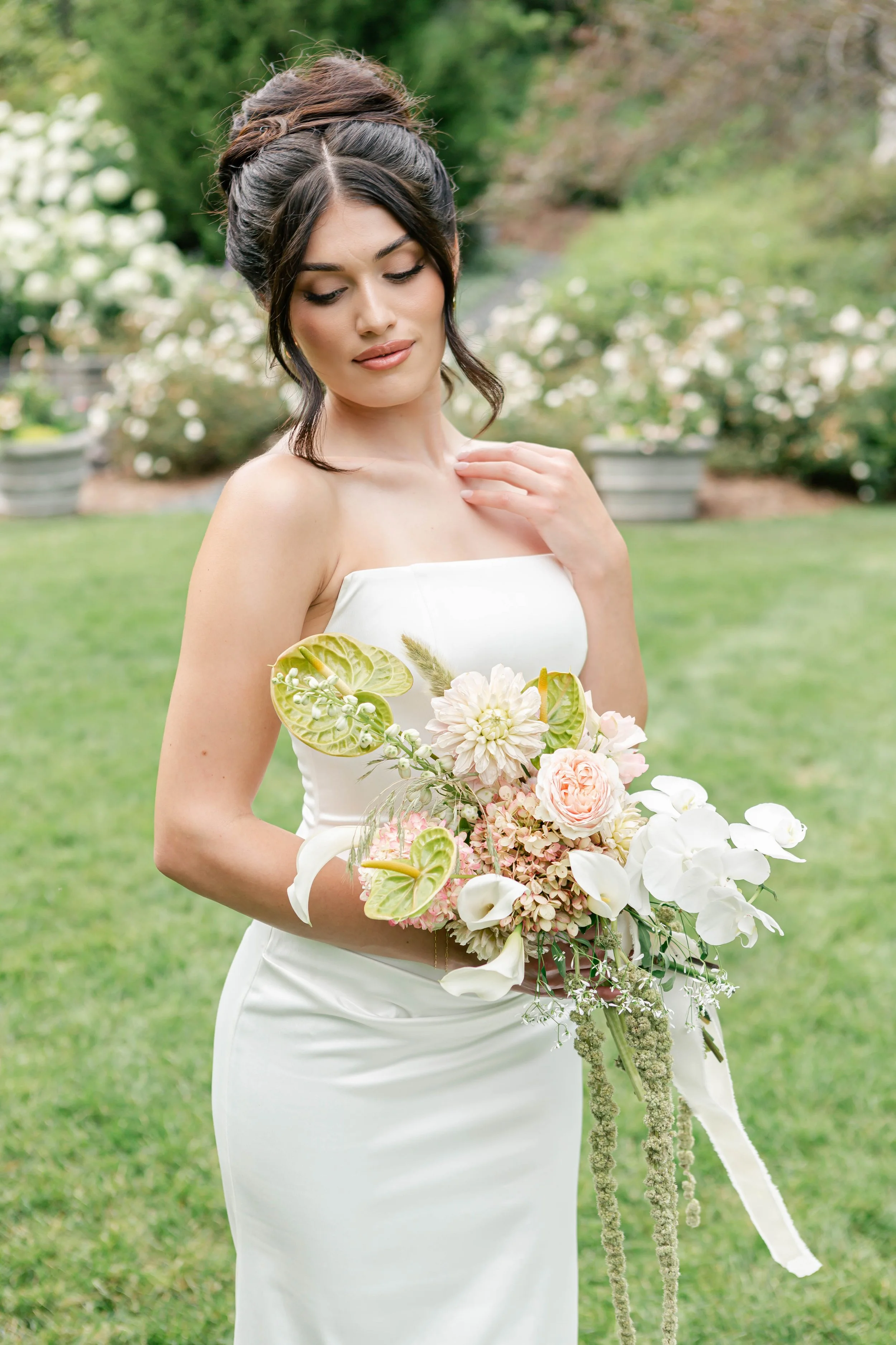A bride with styled dark hair in an outdoor garden setting, holding a bouquet of white and pink flowers including orchids, roses, and greenery, wearing a strapless white wedding dress.