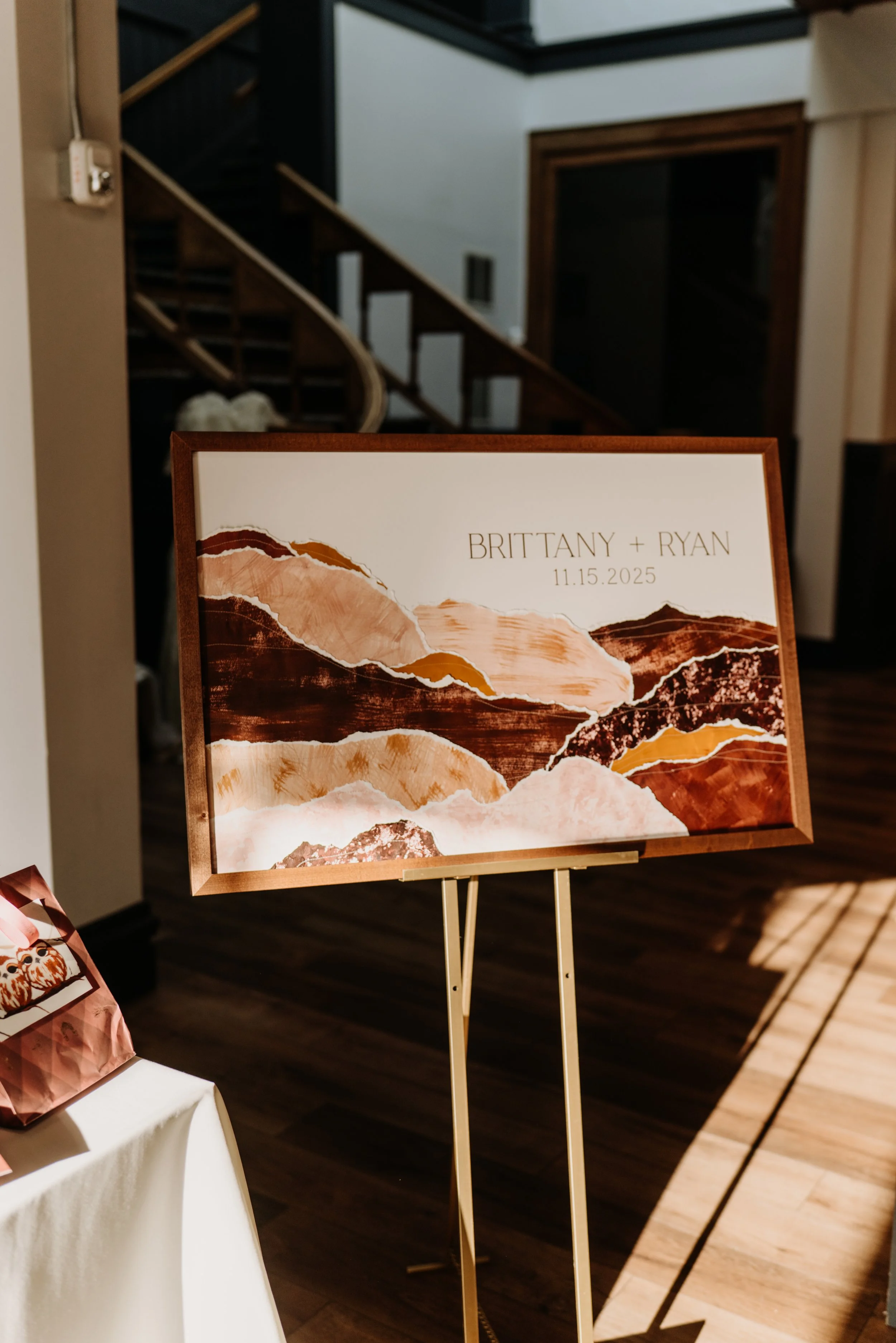 An art display board with a mountain landscape artwork and the names 'Brittany + Ryan' and the date '11.15.2025' on a stand in a room with wooden floors and stairs in the background.