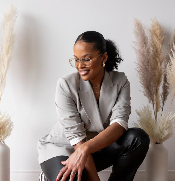 A woman sitting on a white surface, smiling, in a modern, minimalistic room with pampas grass arrangements.