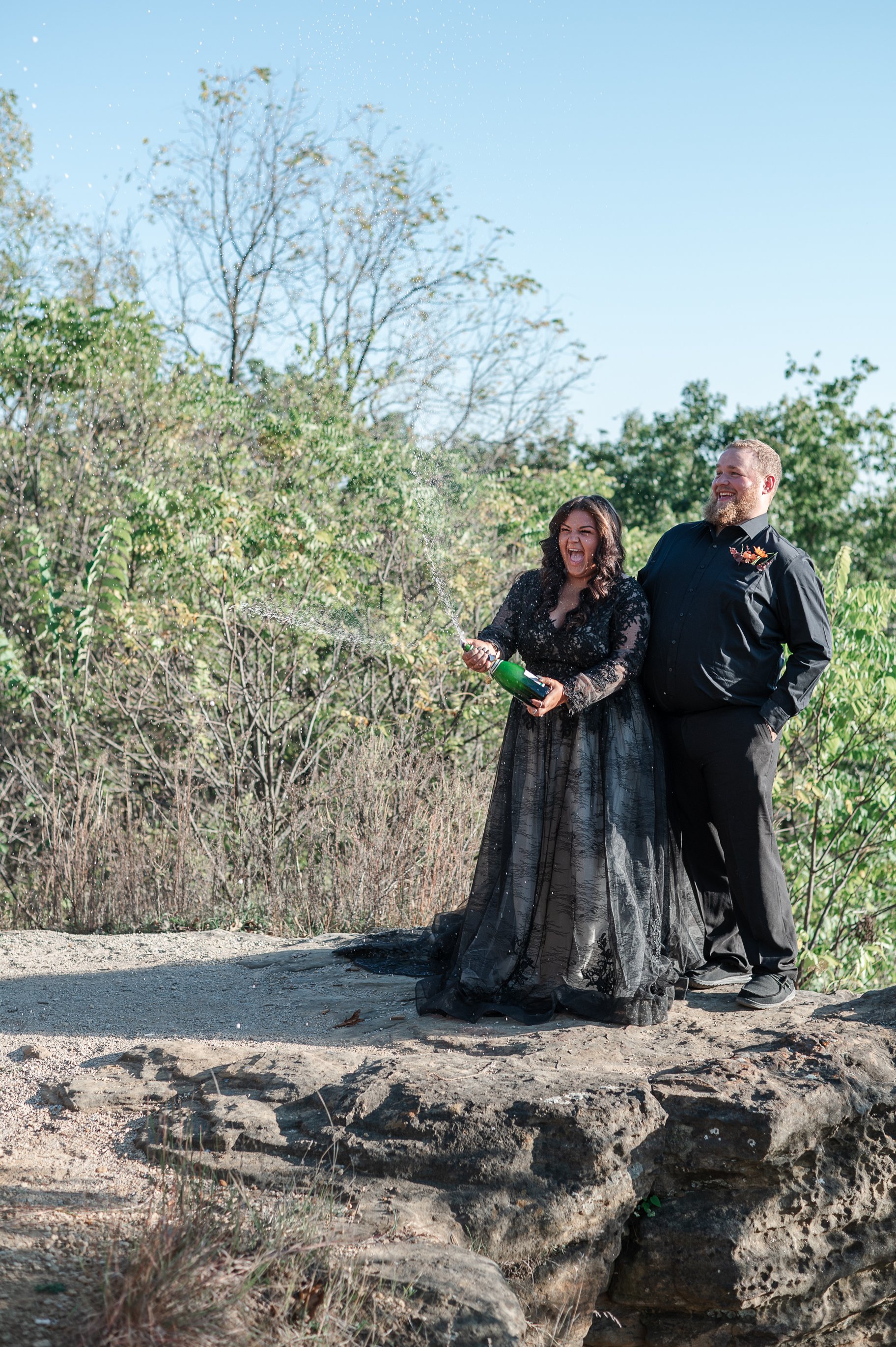 A couple celebrating outdoors on a sunny day, with the woman opening a bottle of champagne, causing it to spray, while both stand on a rocky ledge surrounded by trees, and the man smiles.