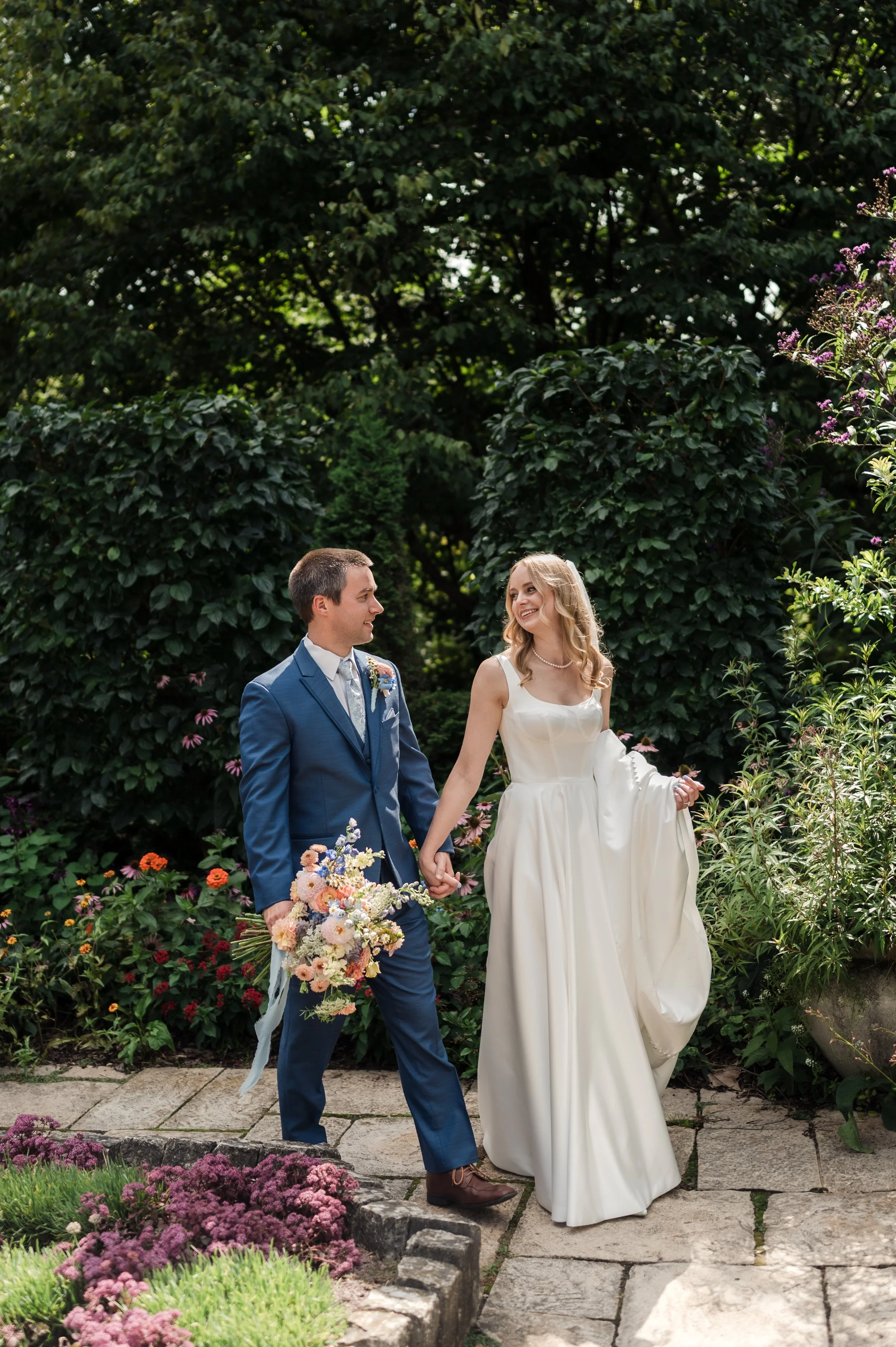 A bride and groom holding hands and smiling in a garden, the bride holding her dress with the other hand, surrounded by flowers and greenery.