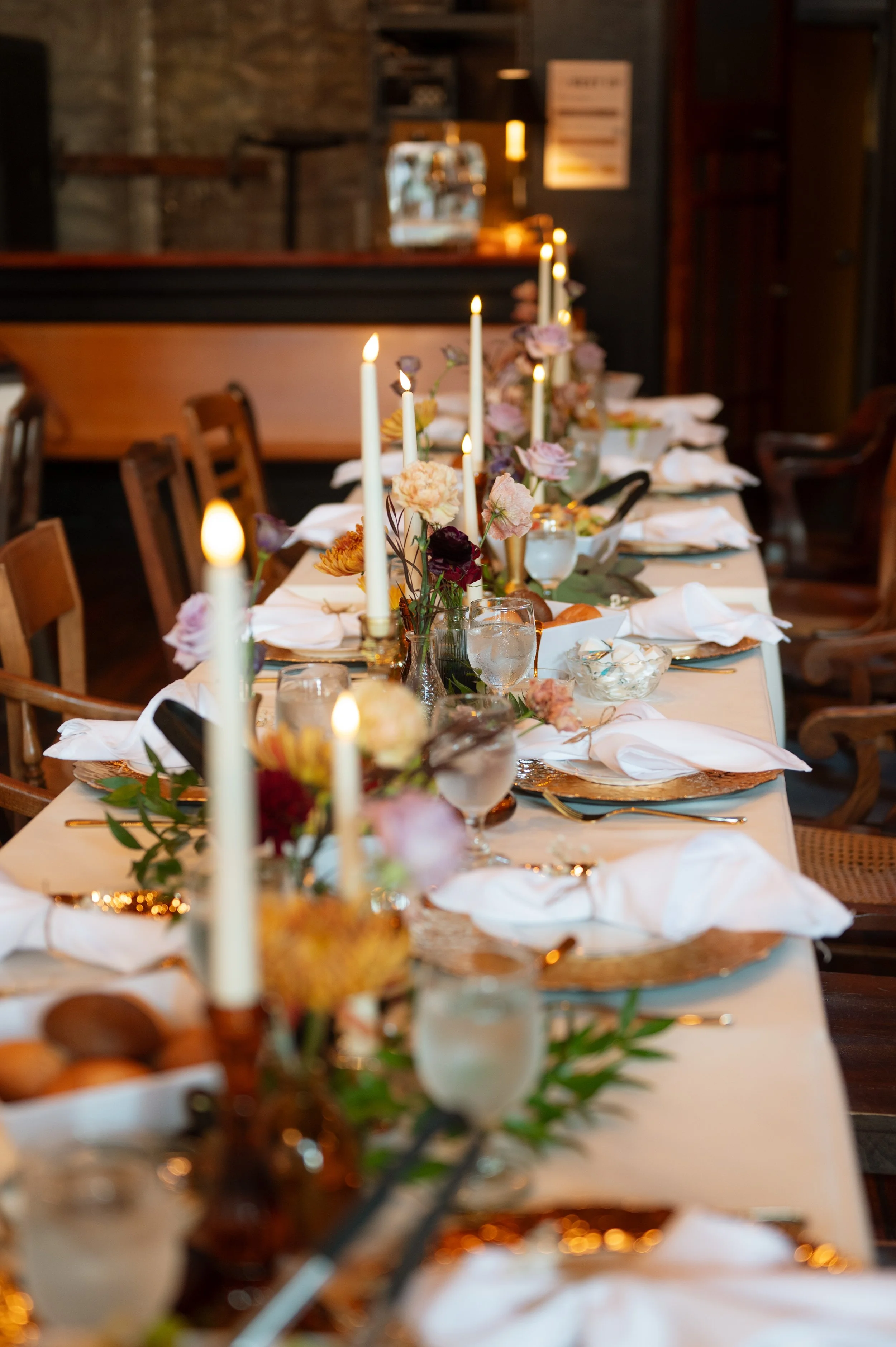 A long dinner table set with white napkins, gold chargers, glassware, and floral centerpieces with white and pink flowers and lit candles in a dimly lit room.