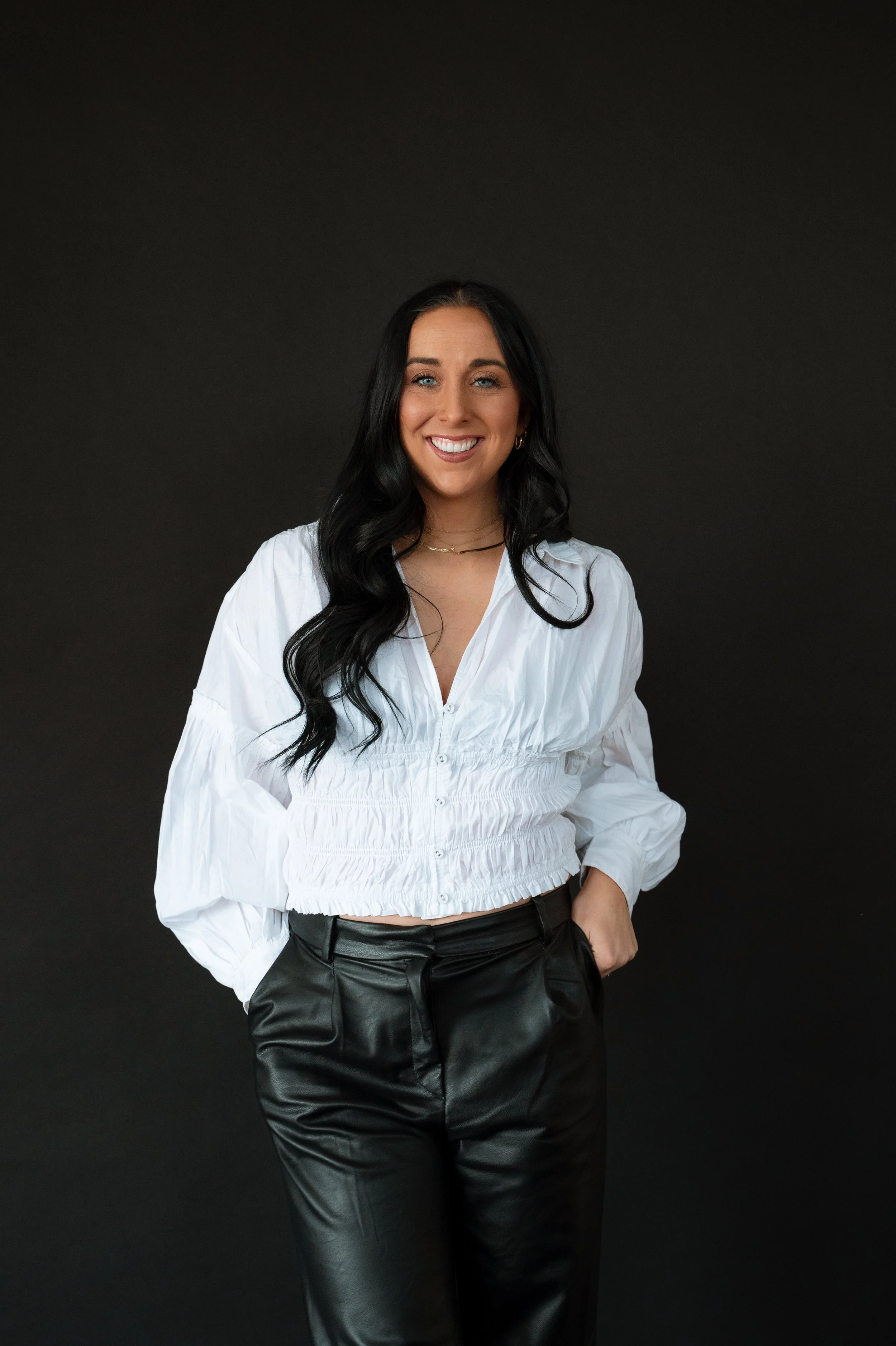A woman with long black hair, blue eyes, and a bright white smile, wearing a white long-sleeve blouse and black leather pants, standing against a dark background.