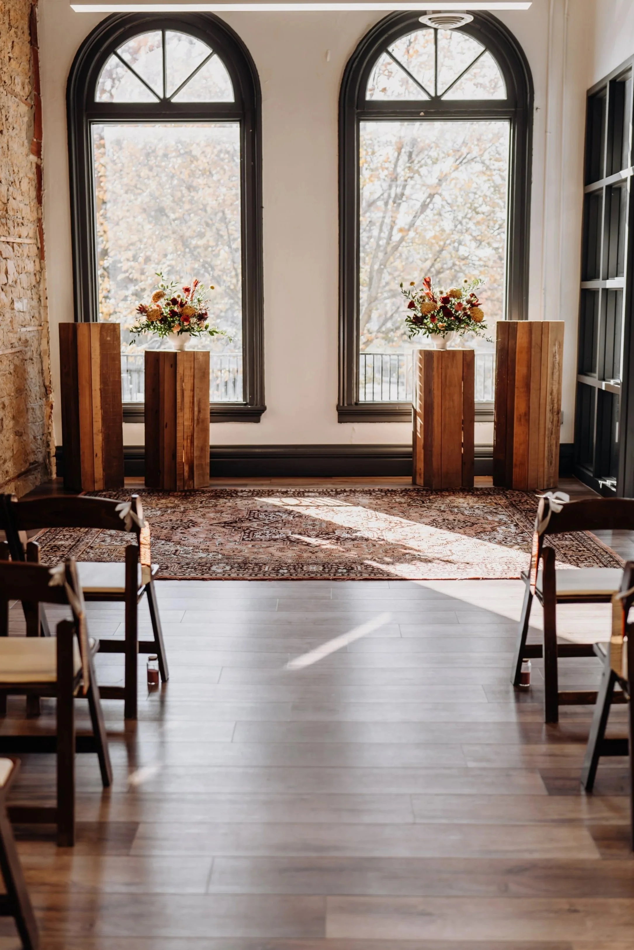 An interior space with two large arched windows overlooking fall foliage, with flower arrangements on wooden pedestals and a patterned area rug on wooden flooring.