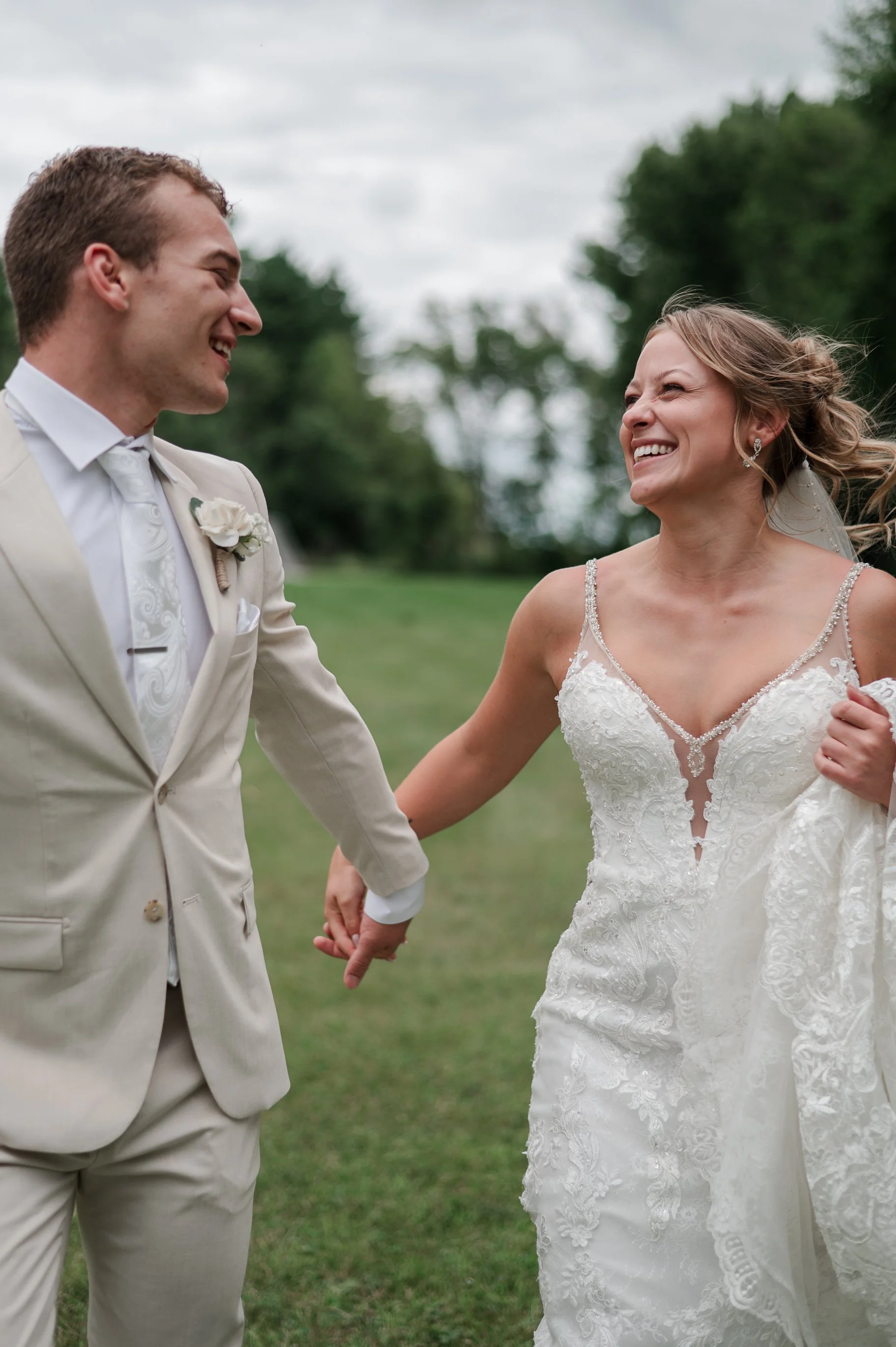A smiling bride and groom holding hands outdoors on a cloudy day.