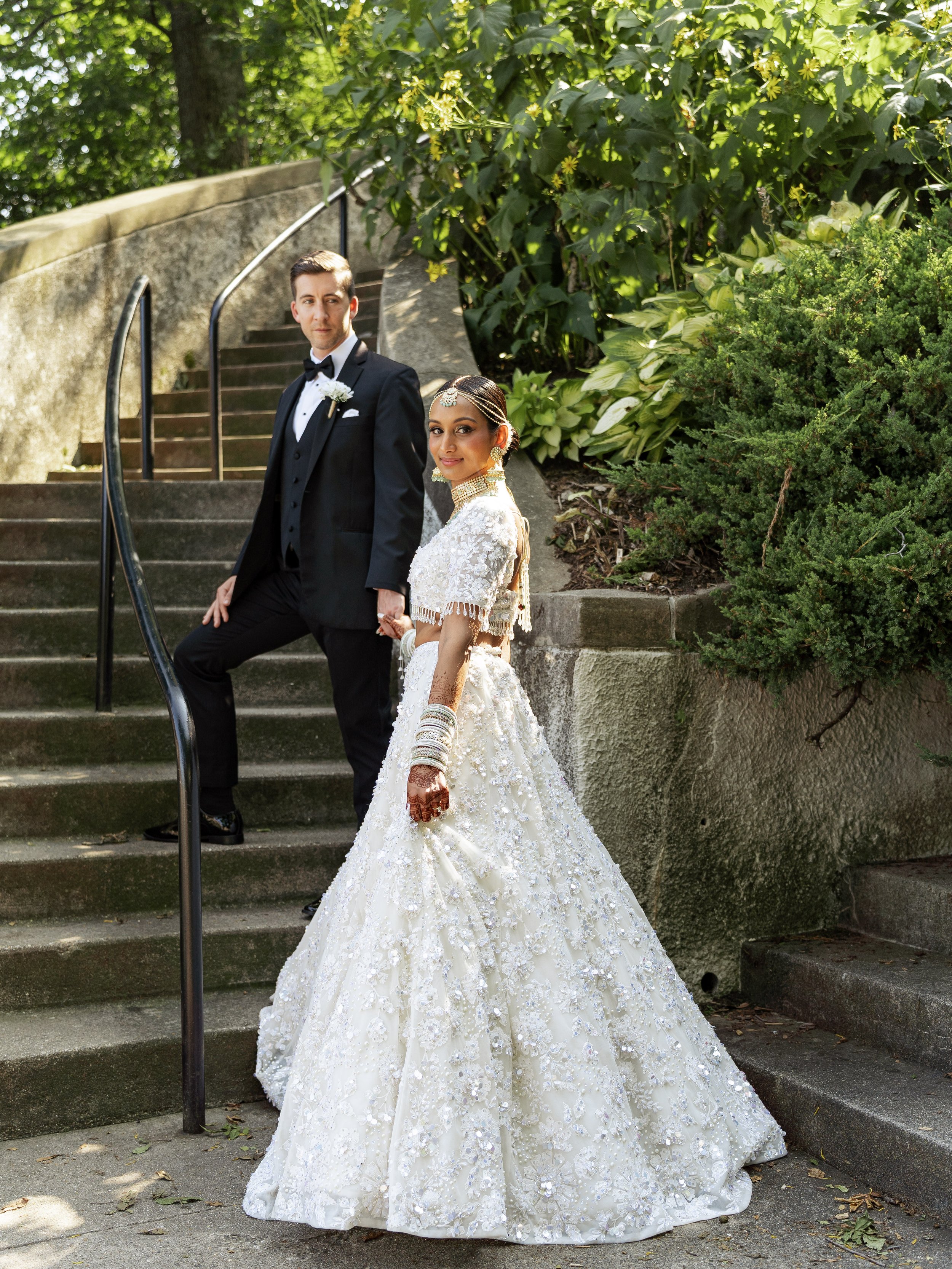 A bride and groom in wedding attire standing on outdoor stairs with lush greenery around them.