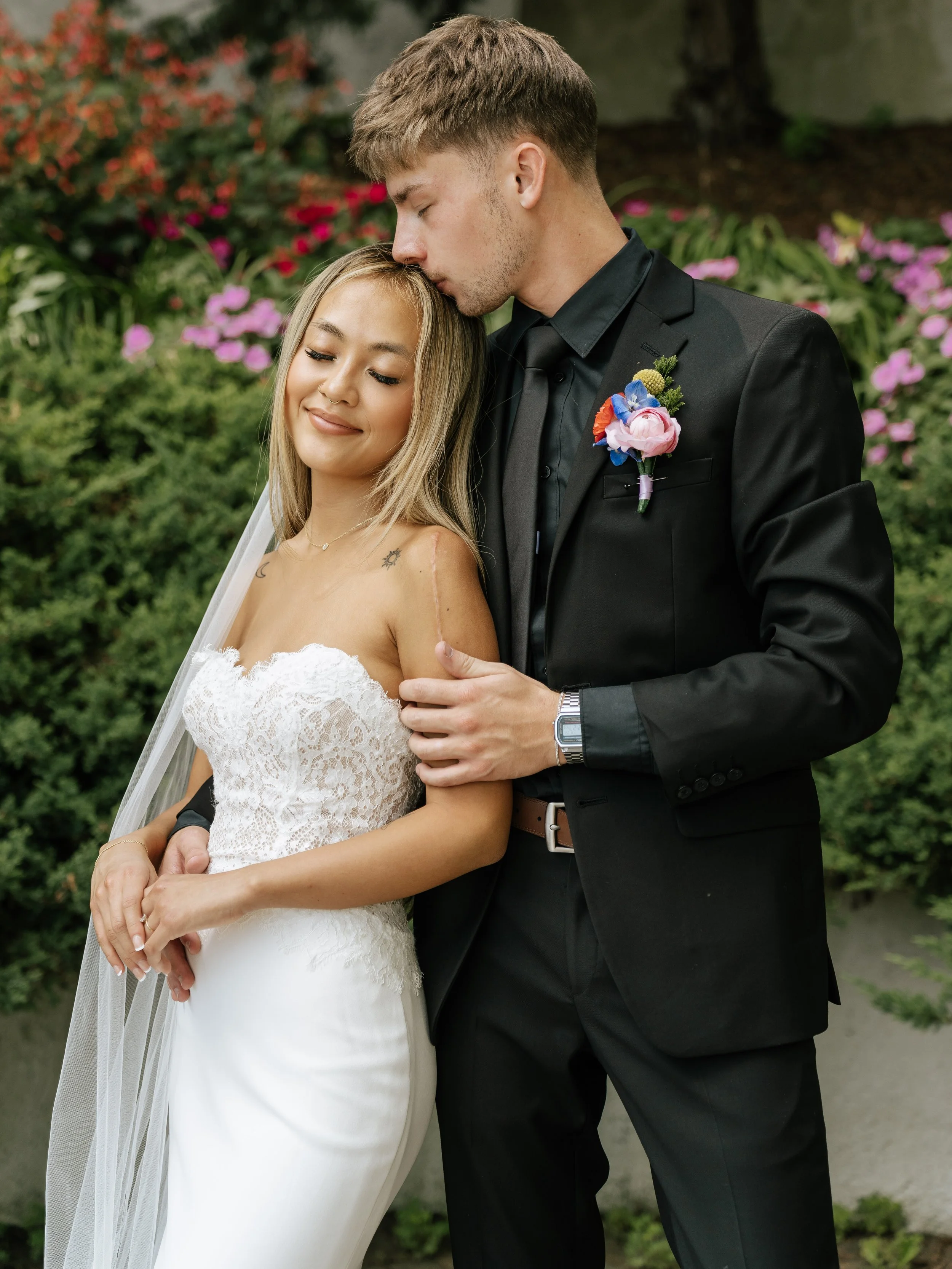 A bride in a white lace wedding gown and a groom in a black suit with a boutonnière, standing outdoors in front of colorful flowers and bushes, with the groom kissing the bride's forehead and the bride smiling with her eyes closed.