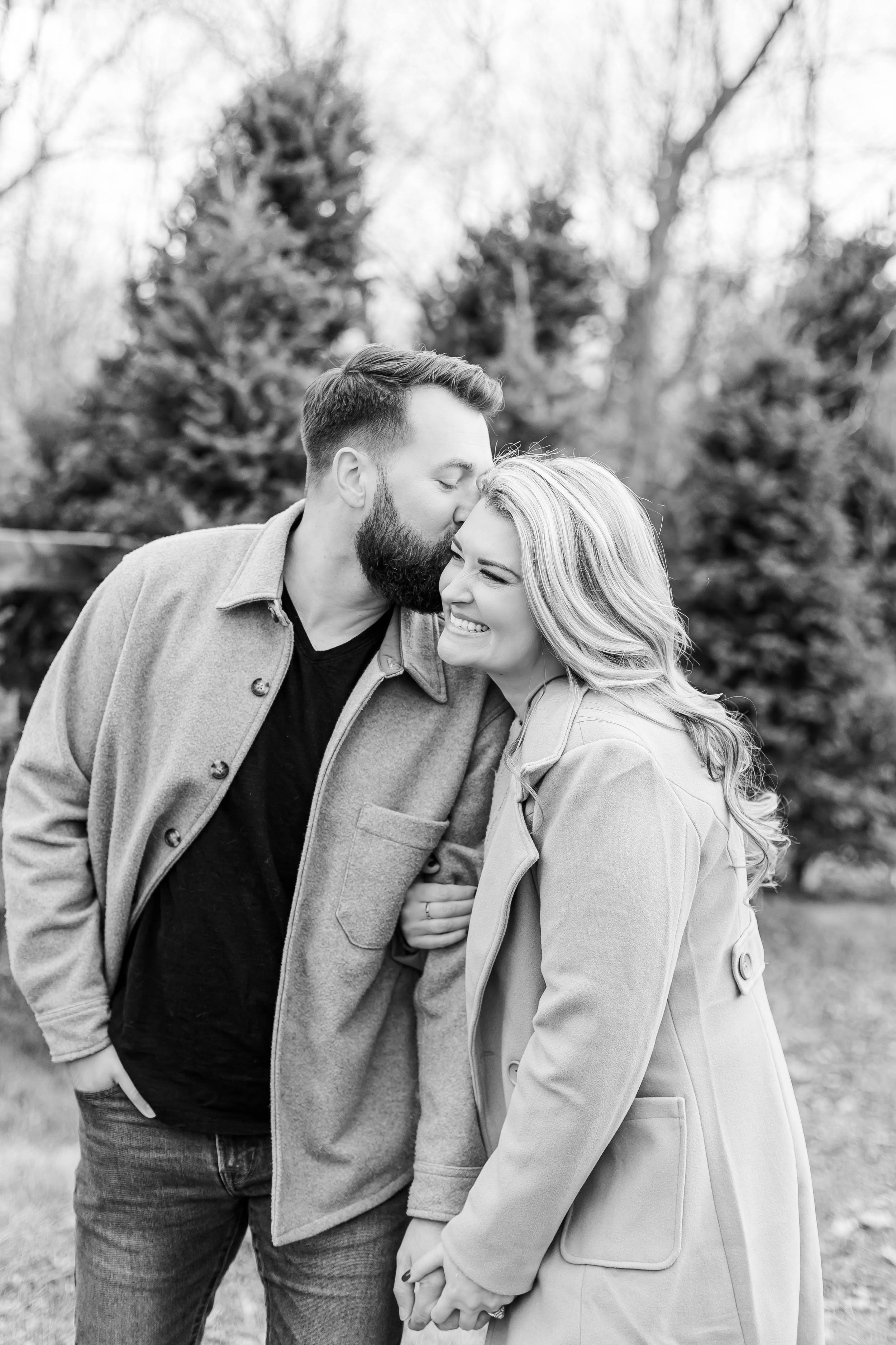 A black-and-white photo of a couple standing outdoors, smiling and holding hands, with trees in the background. The man is kissing the woman's forehead, and she is smiling happily.