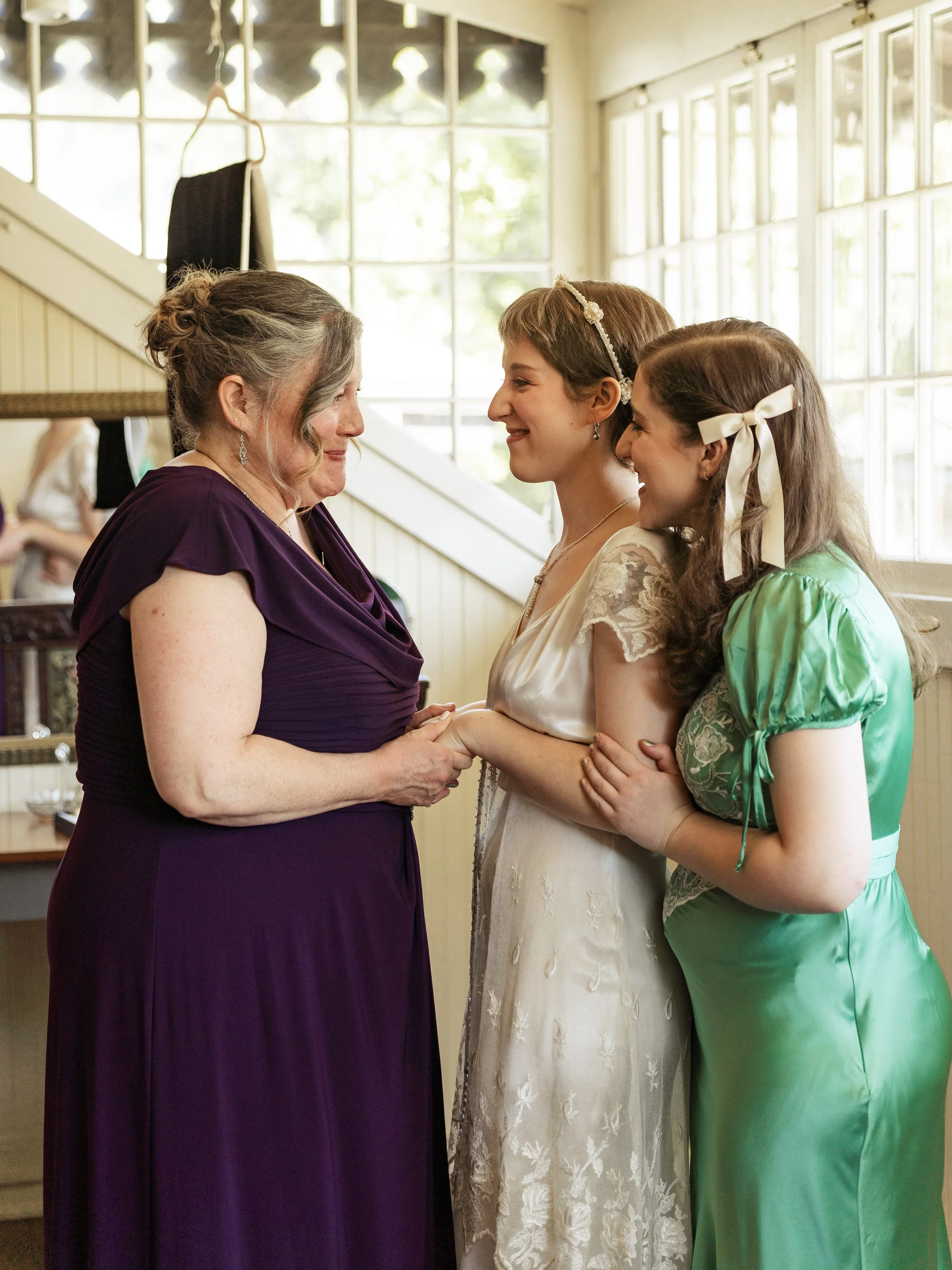 Three women smiling and holding hands inside a sunlit room, with large windows and a staircase in the background.