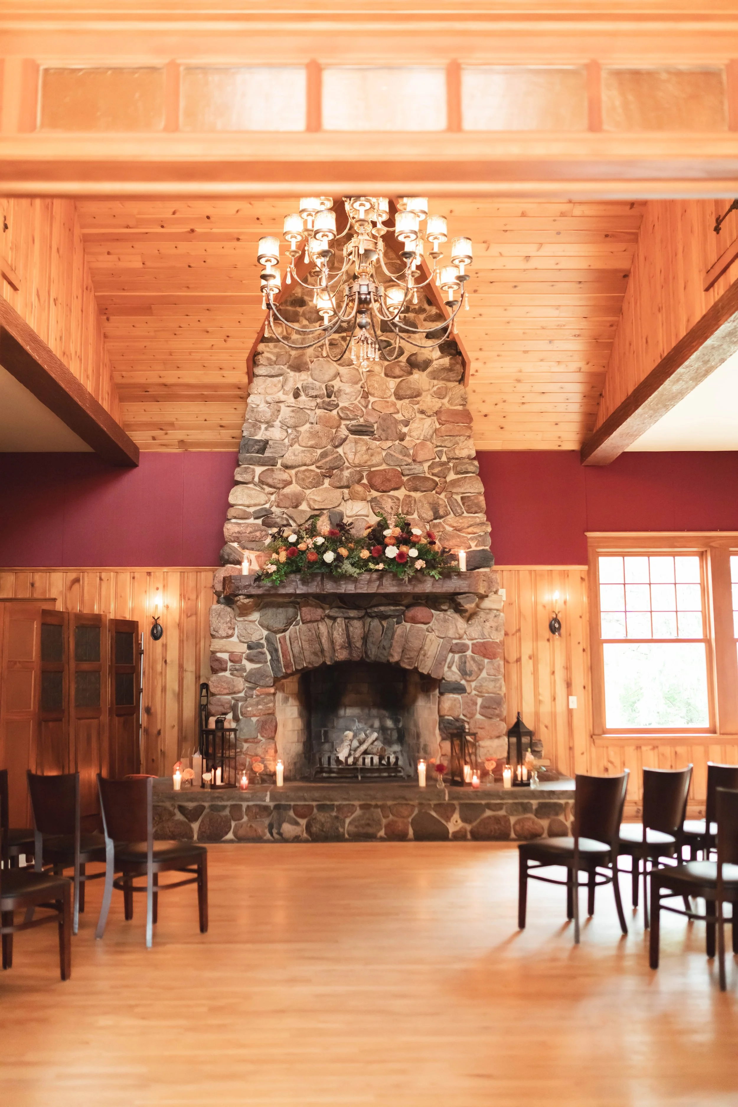 Interior of a lodge or cabin with a large stone fireplace decorated with candles and flowers, wooden walls, and a chandelier hanging from a wooden ceiling.