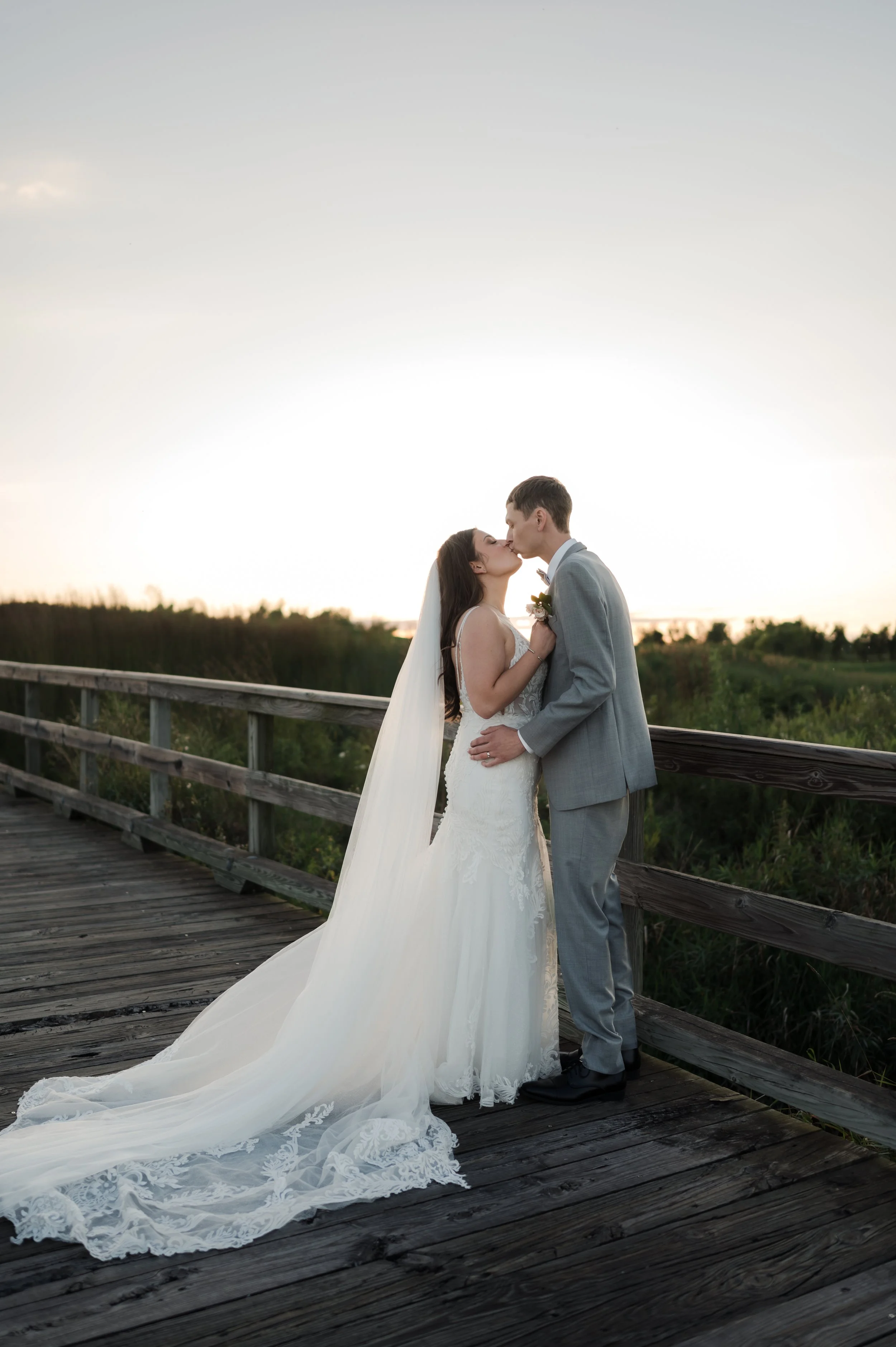 A bride and groom kissing on a wooden bridge at sunset, with the bride in a white wedding gown and veil, and the groom in a gray suit.
