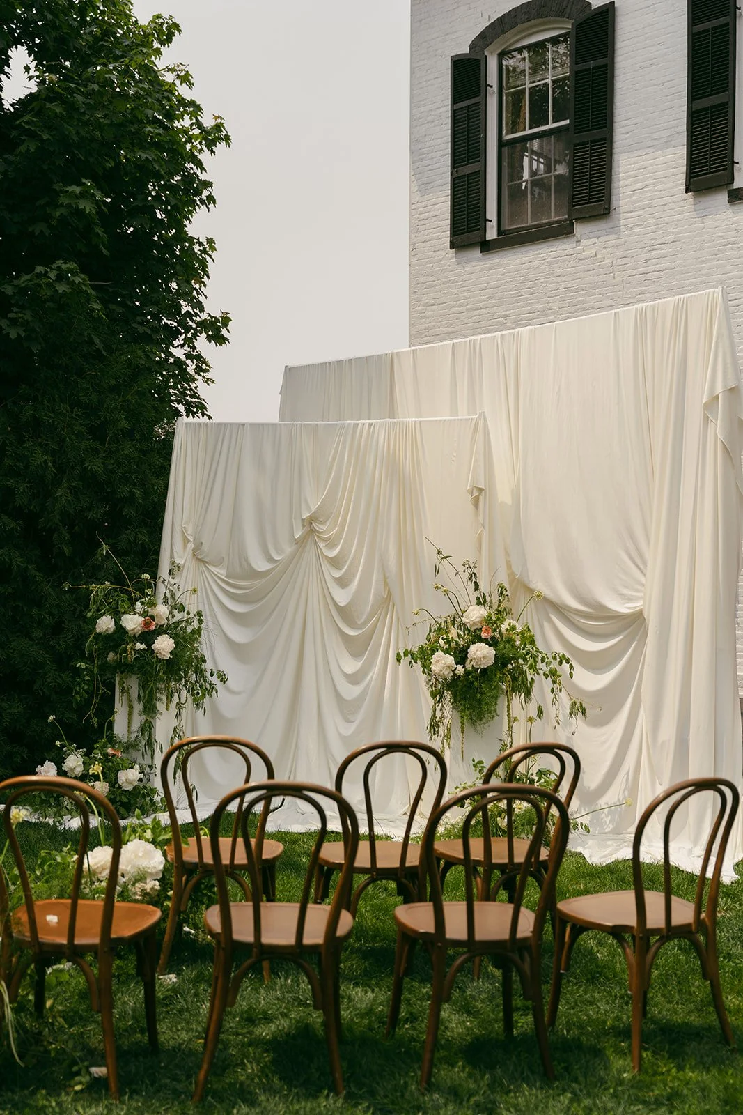 Outdoor wedding setup with a cream draped backdrop and floral arrangements, wooden chairs on grass, behind a white brick building with black shutters.