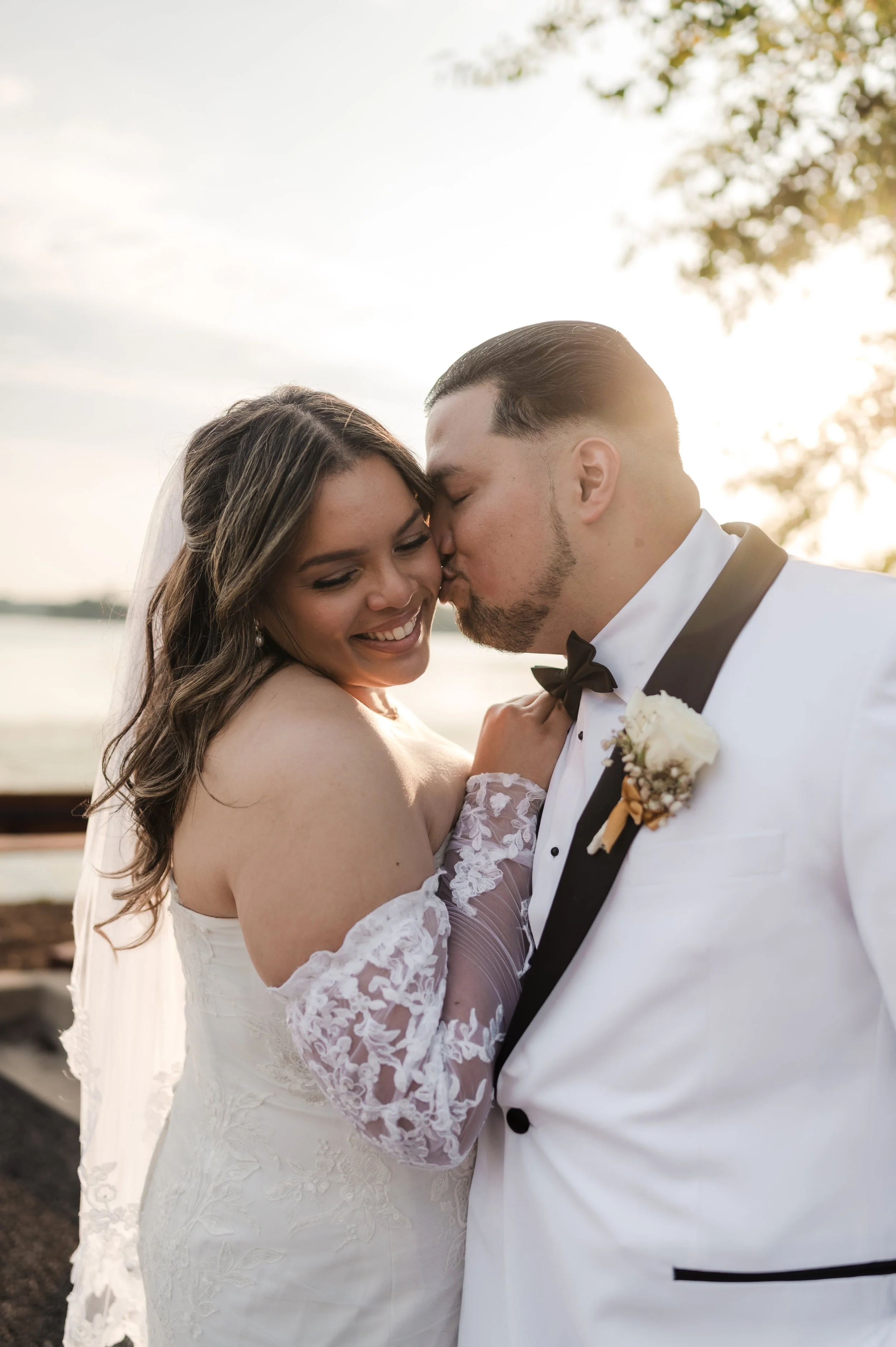 A newlywed couple sharing a tender moment outdoors at sunset, with the groom kissing the bride on the forehead. The bride is smiling with eyes closed, dressed in a white wedding gown with lace sleeves, and the groom wears a white tuxedo with a black 