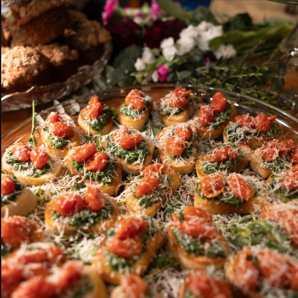 Close-up of a platter of bruschetta topped with tomato, basil, and shredded cheese, on a wooden table with flowers in the background.
