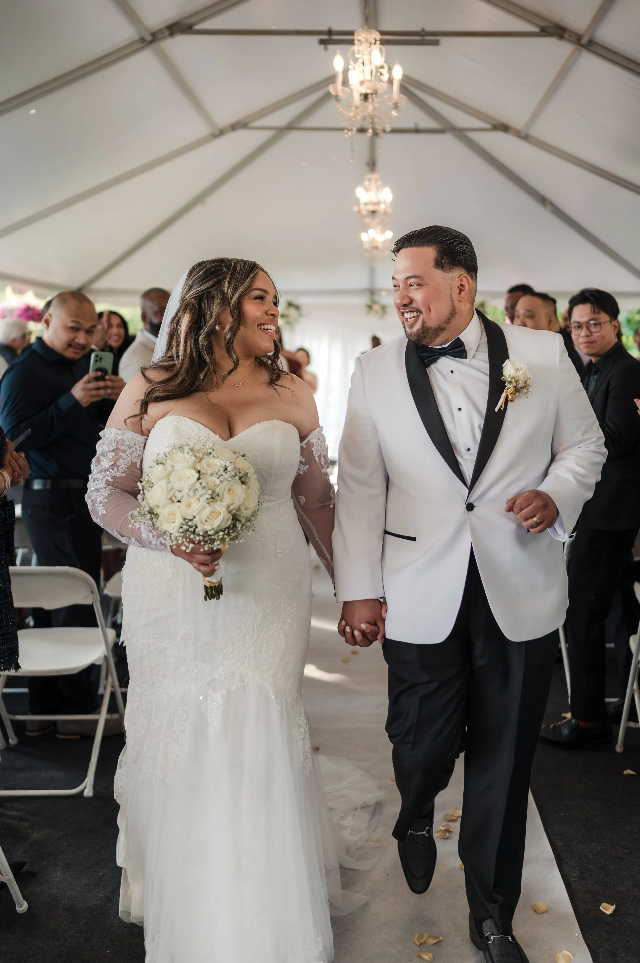 A newlywed couple holding hands and smiling at each other during their wedding ceremony, with guests and chandeliers in the background.