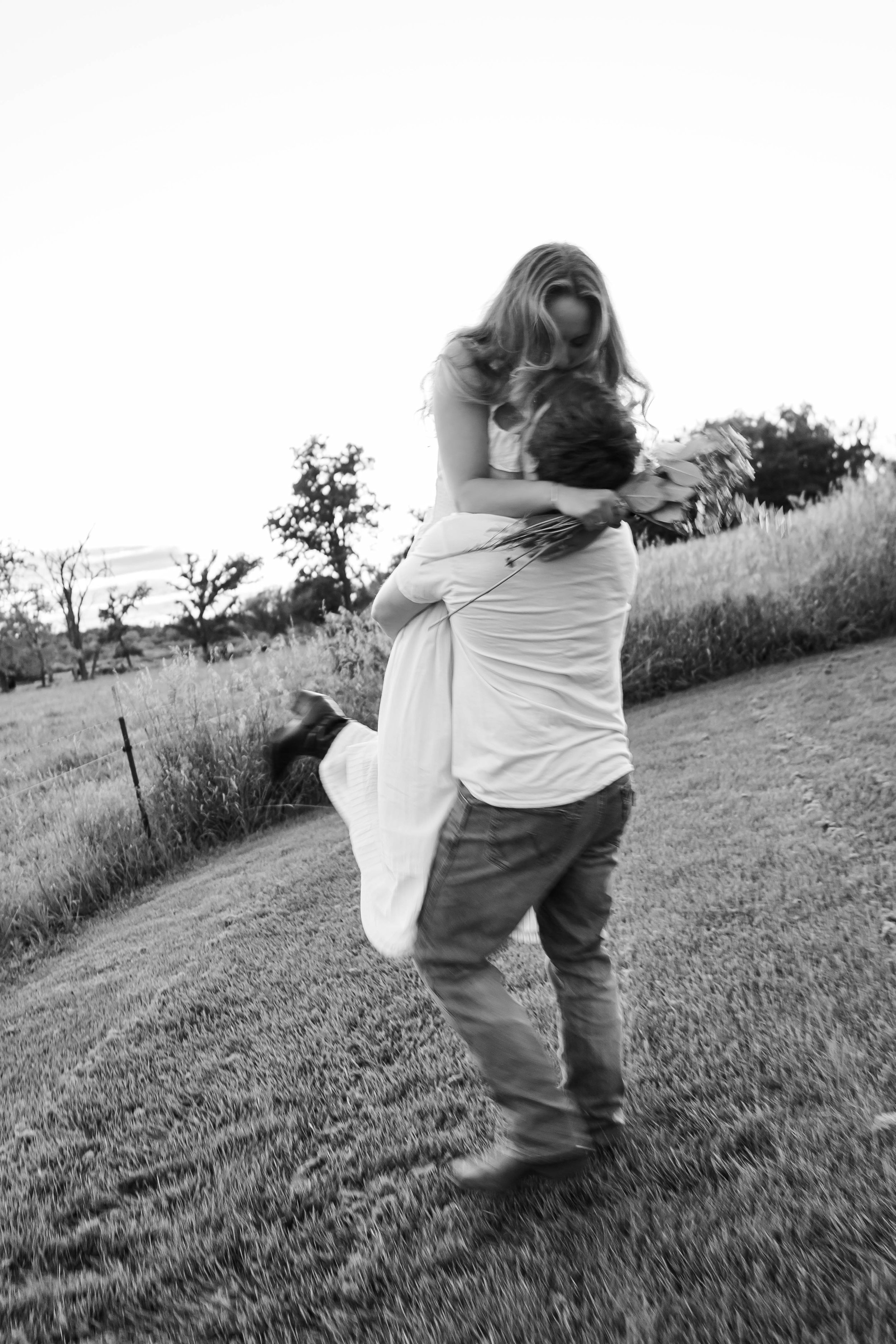 A black-and-white photo of a man lifting a woman in a field, with trees in the background, and the woman holding a bouquet of flowers.