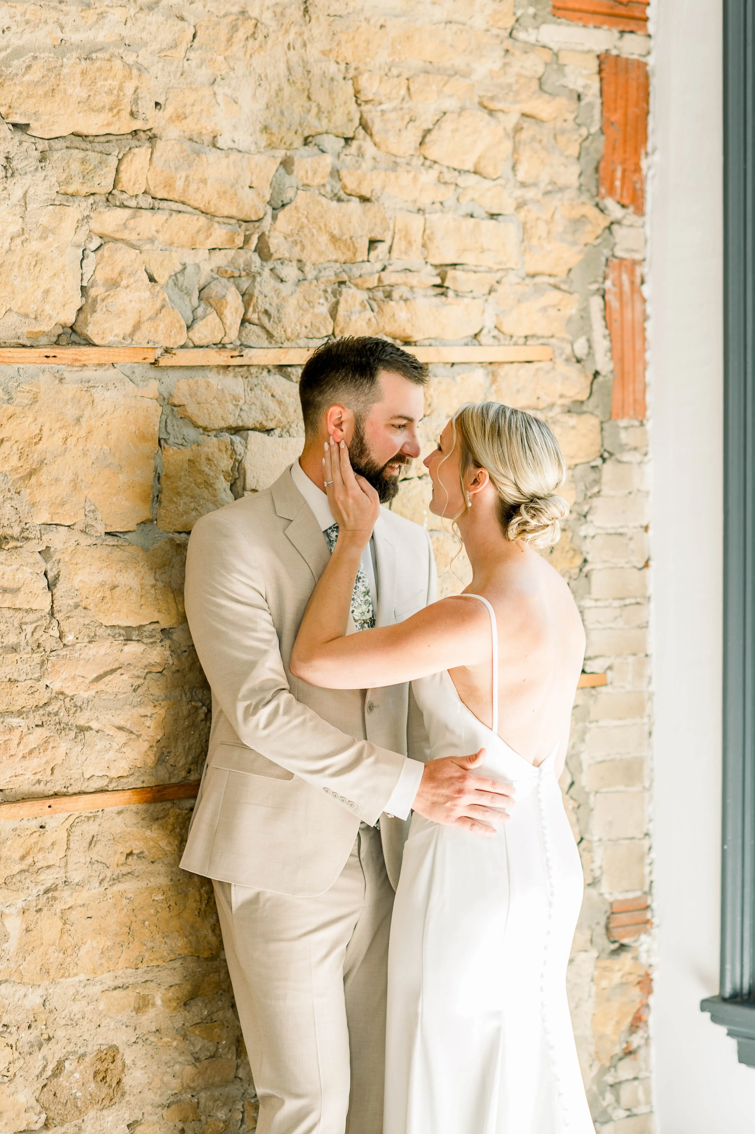 A wedding couple standing close together by a textured stone wall, embracing and gazing into each other's eyes, with a large window on the right.