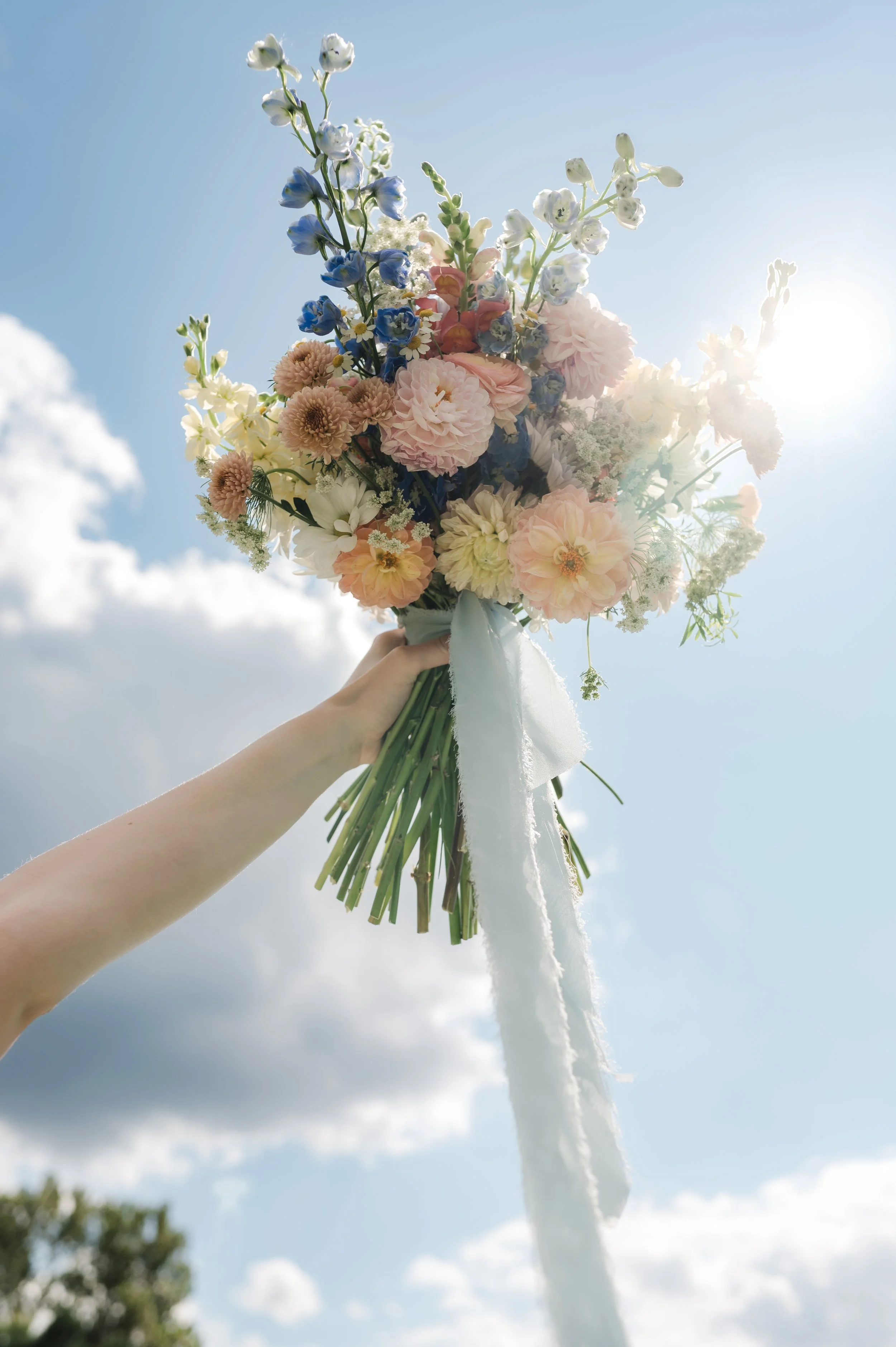 A person holding a bouquet of colorful flowers against a blue sky with clouds and sunlight.