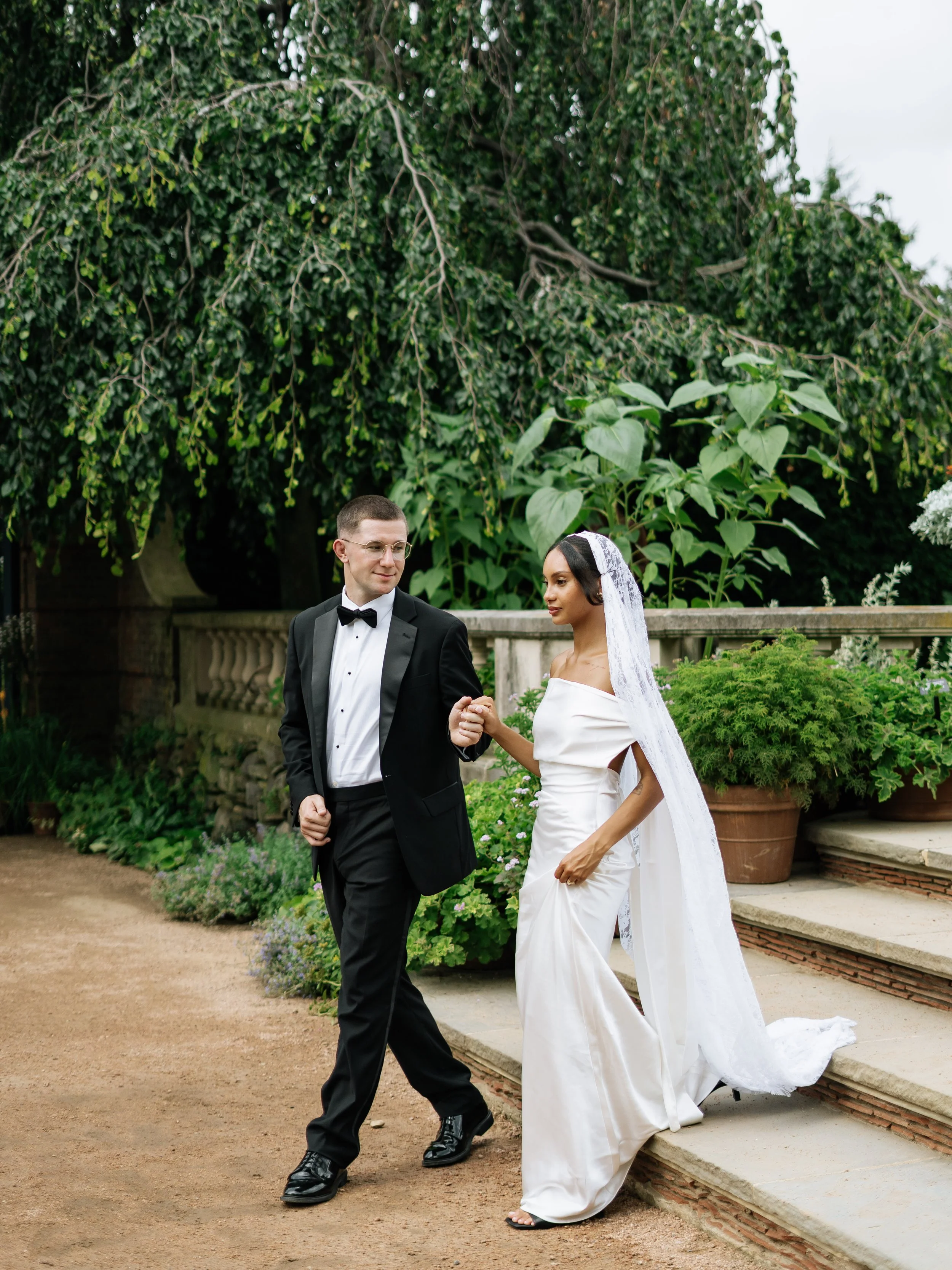 A groom in a black tuxedo helping a bride in a white wedding dress descend steps in a garden setting with lush greenery and potted plants.