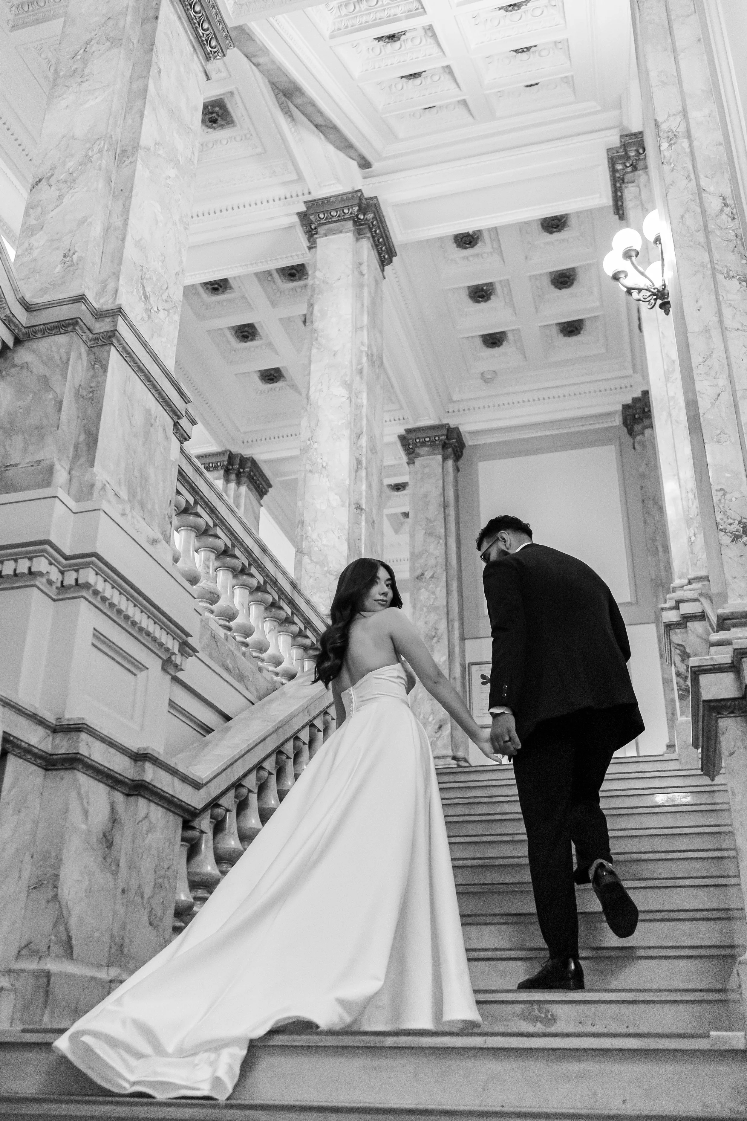 A black and white photo of a bride and groom walking up marble stairs in an ornate interior with high ceiling and columns. The bride wears a strapless wedding gown, and the groom wears a dark suit.