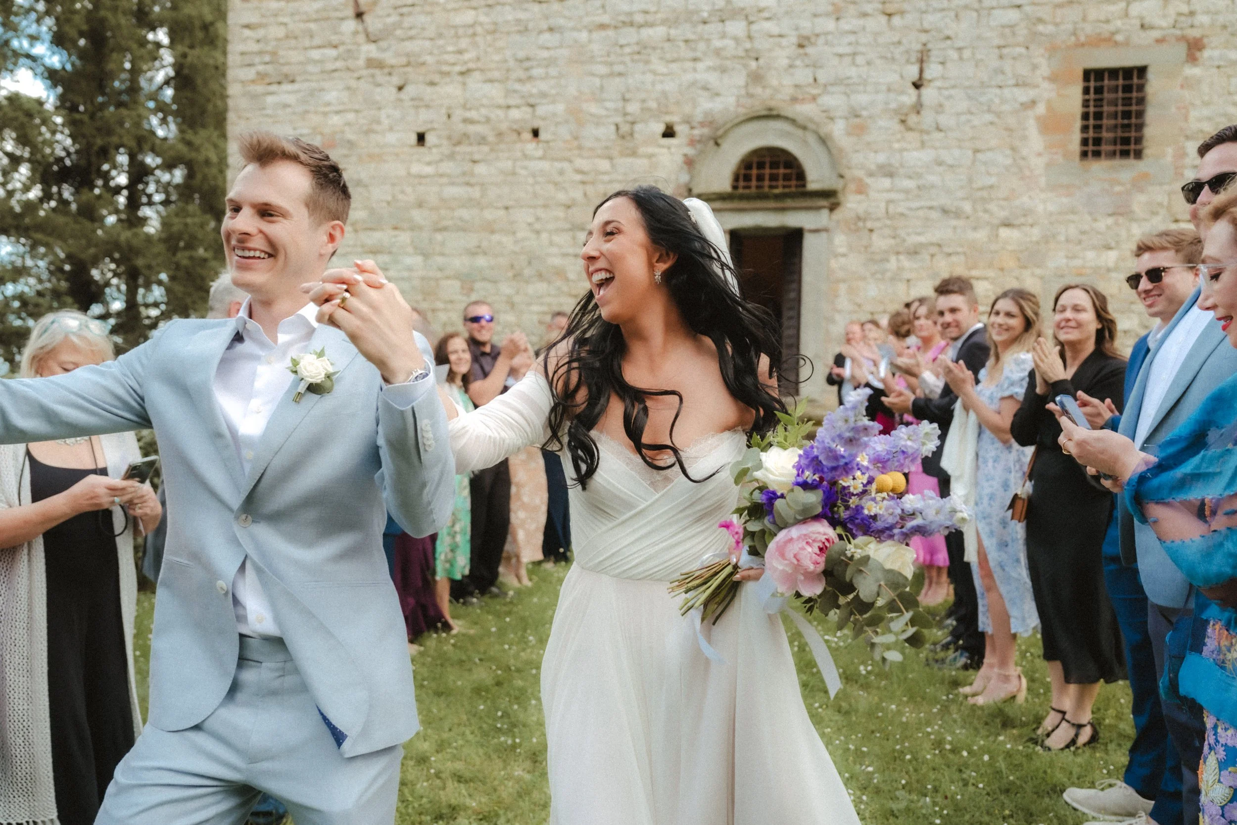A wedding celebration outdoors with a bride and groom dancing, surrounded by guests clapping and smiling, in front of an old stone building.