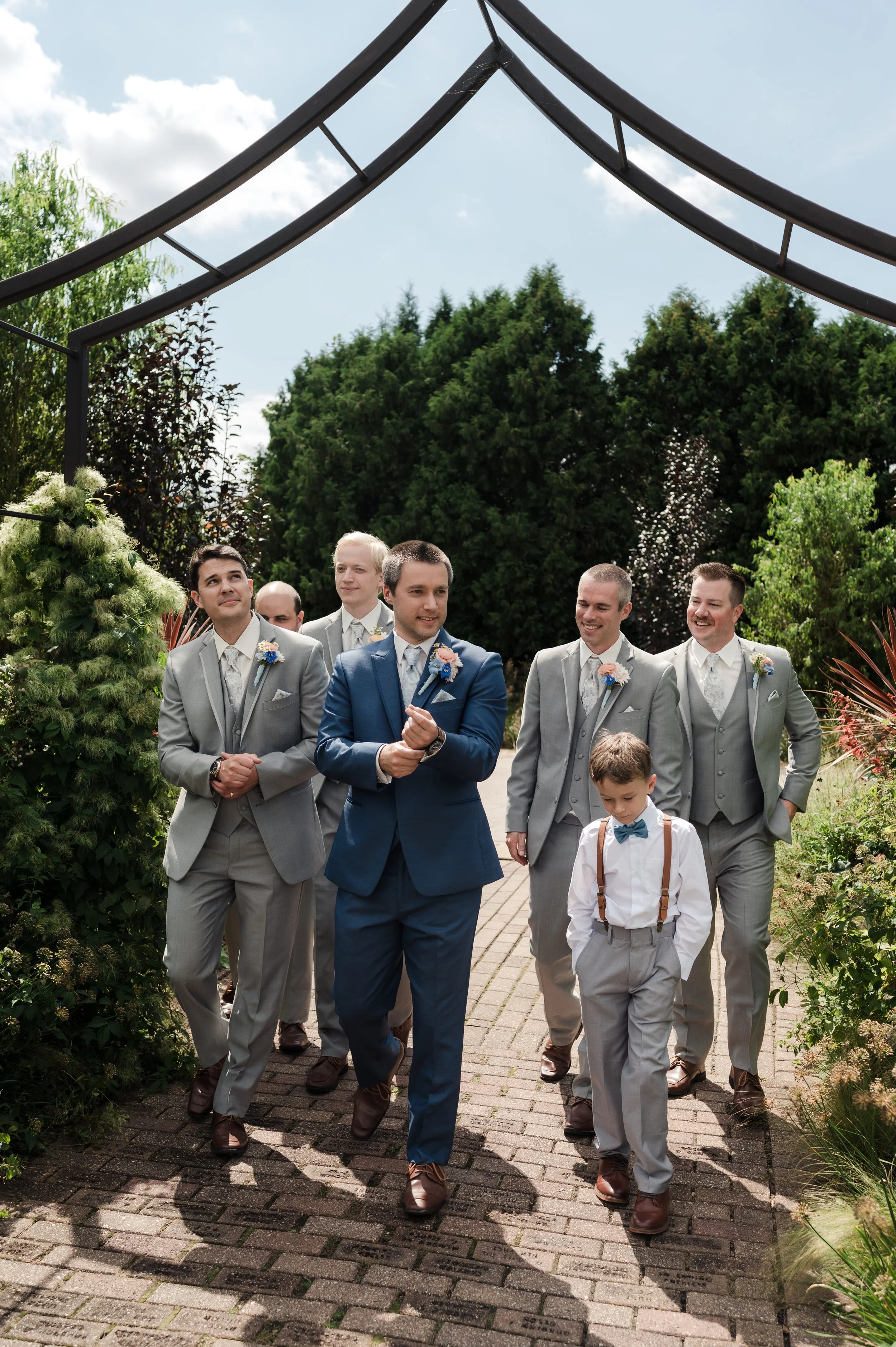 A group of men dressed in suits walking outdoors on a paved path, with lush greenery and flowering plants surrounding them on a sunny day.