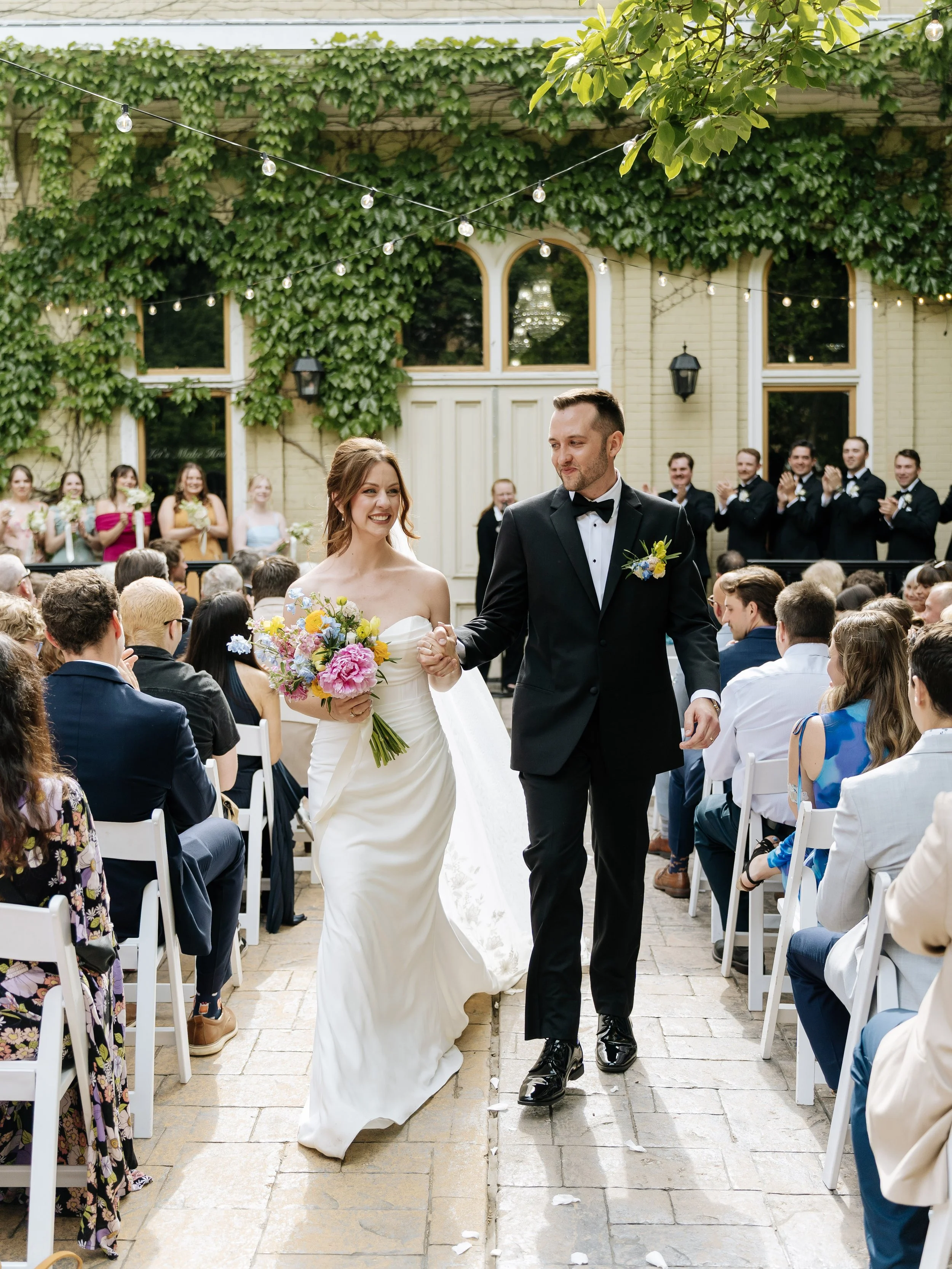 A bride and groom walking down the aisle at their outdoor wedding ceremony, smiling and holding hands. Guests seated on white chairs are clapping, and bridesmaids are visible in the background. The setting is decorated with string lights and green fo