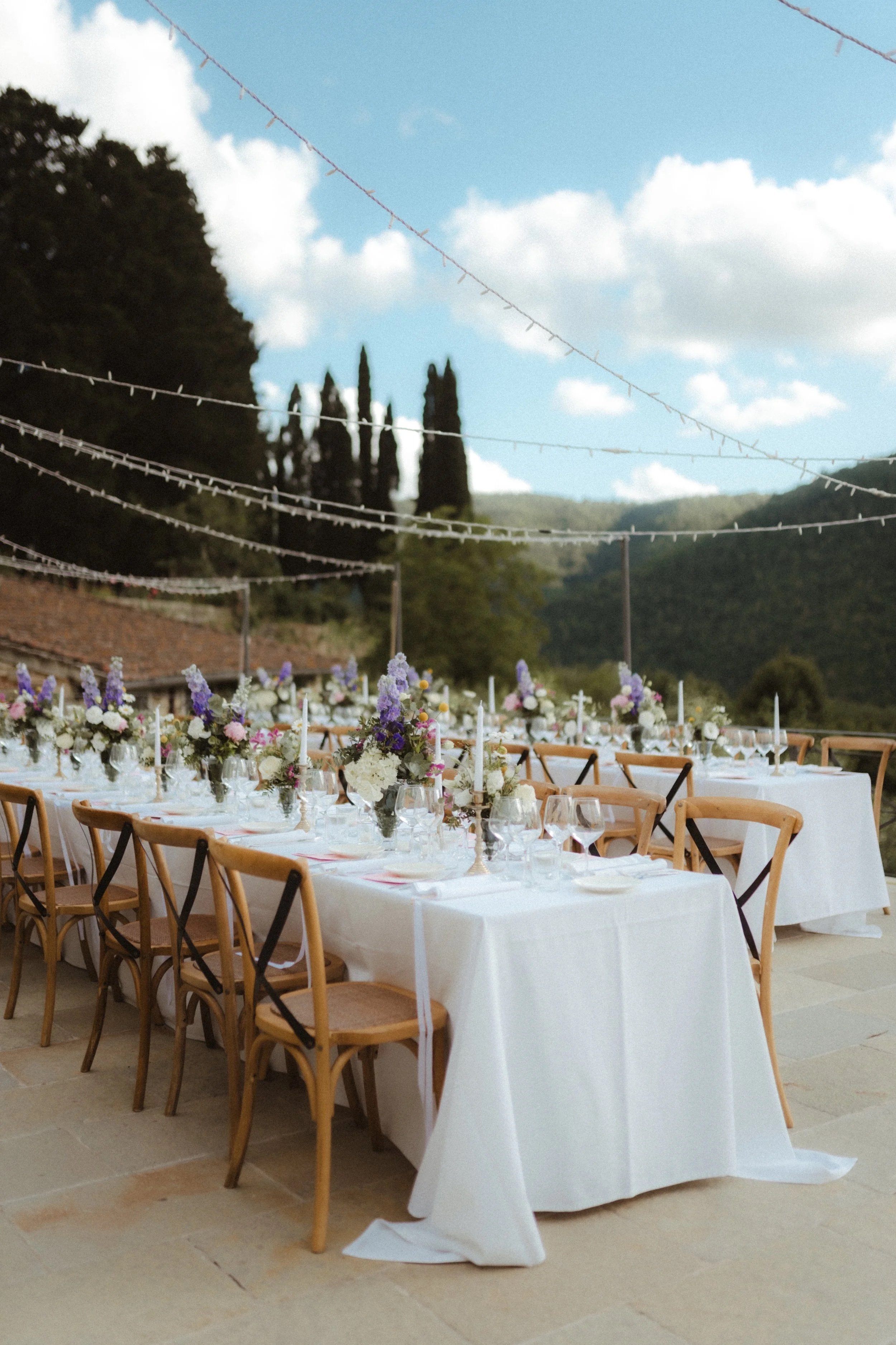 Long reception table with white tablecloth, floral centerpieces, and place settings, outdoor setting with string lights and a scenic backdrop of mountains and trees.