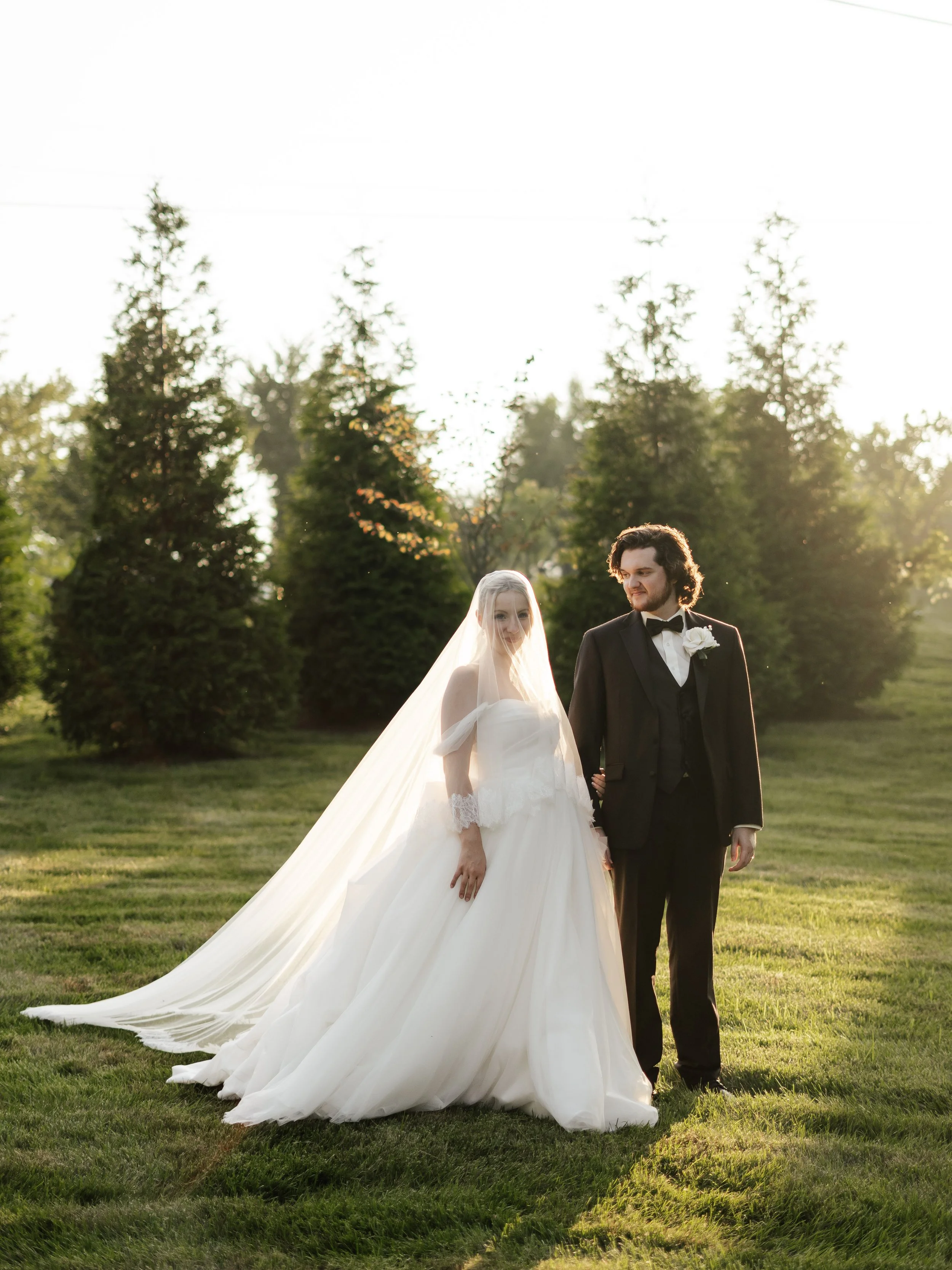 A bride and groom standing outdoors on grass with tall trees in the background, dressed in wedding attire, with the bride in a white gown and veil, and the groom in a black tuxedo, during sunset.
