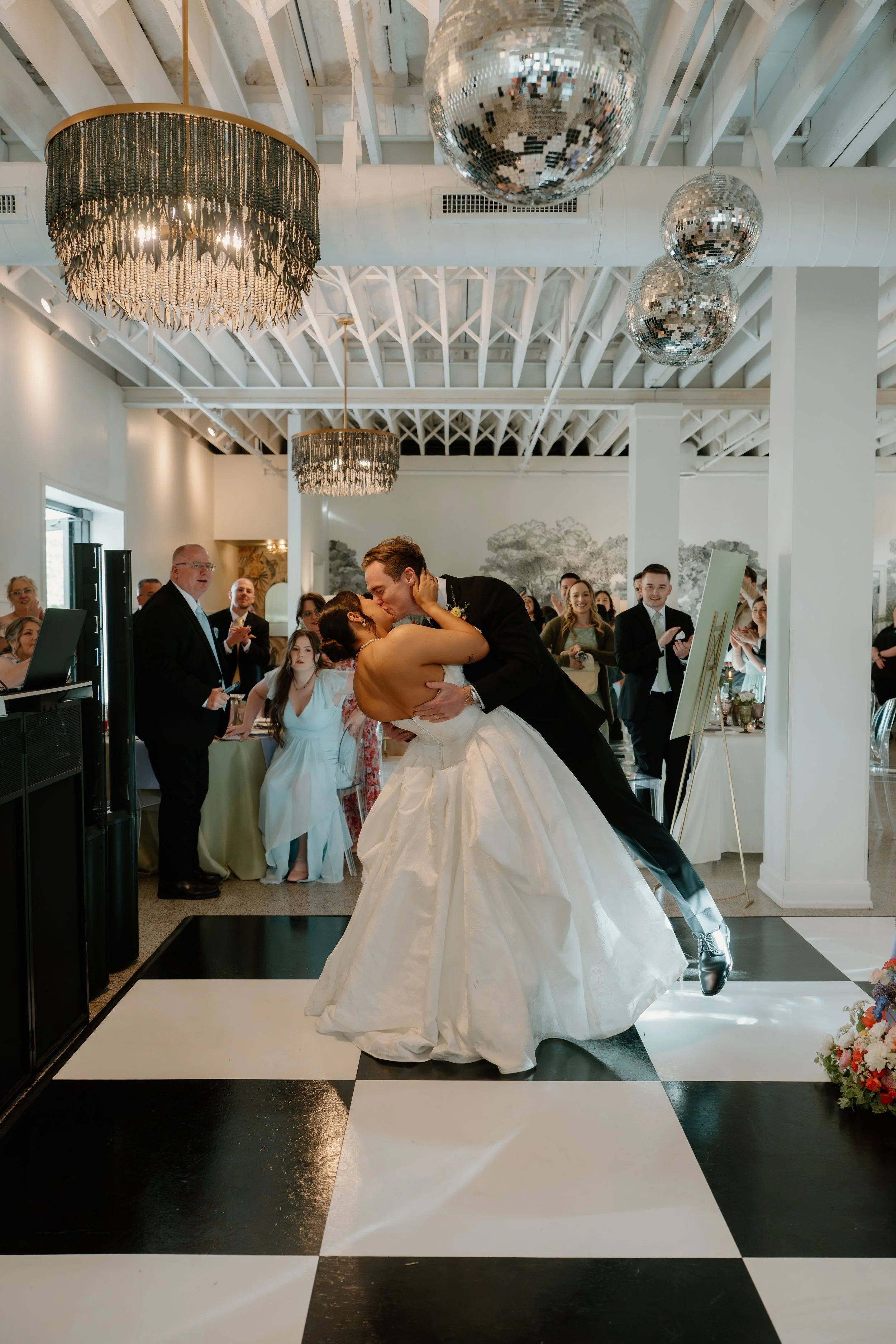 A bride and groom share a dance at their wedding reception, with guests watching and celebrating in the background, inside a decorated venue with chandeliers and disco balls.