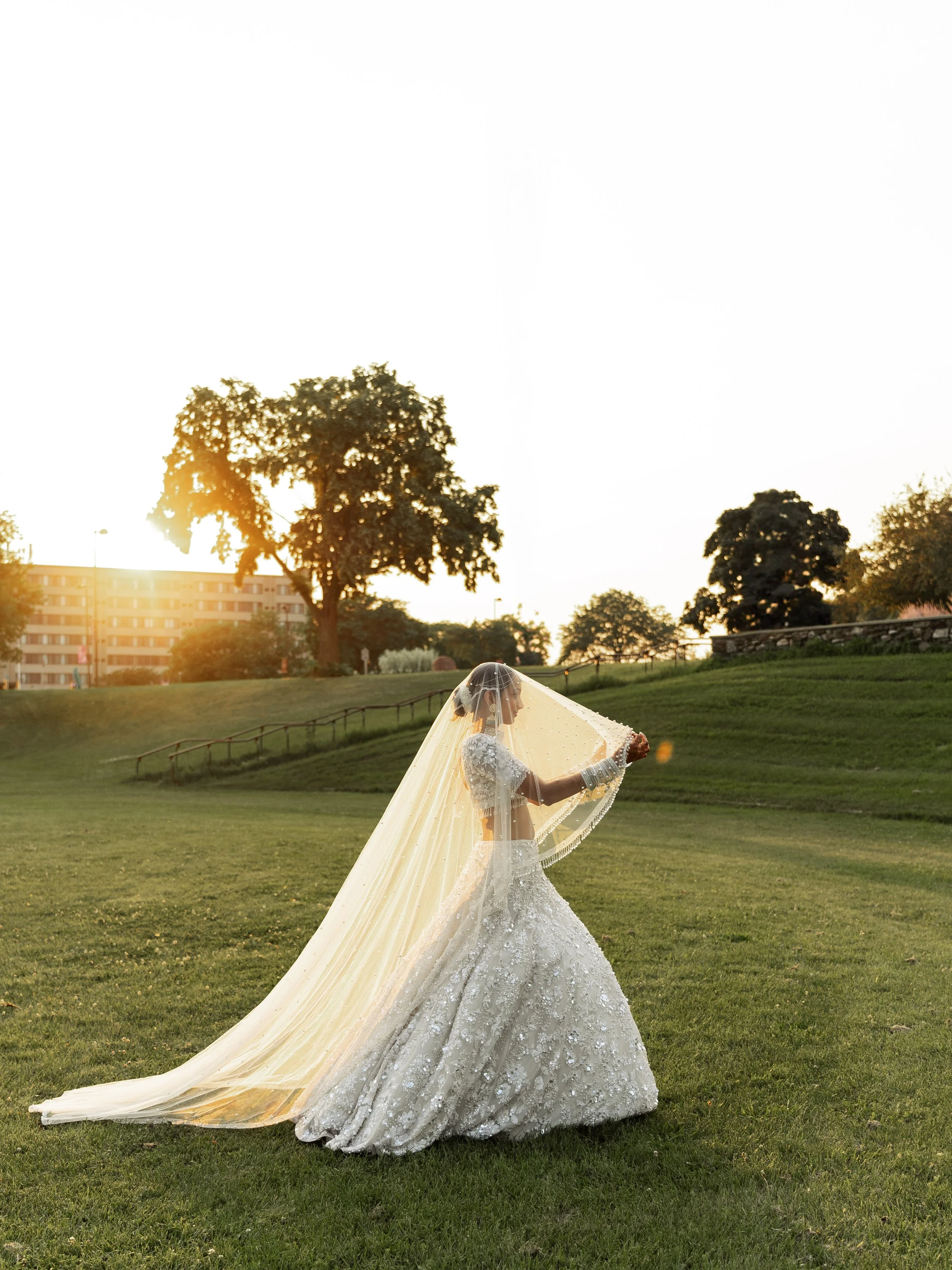 Bride wearing a traditional white wedding gown and veil, standing on a grassy field during sunset, with trees and a building in the background.