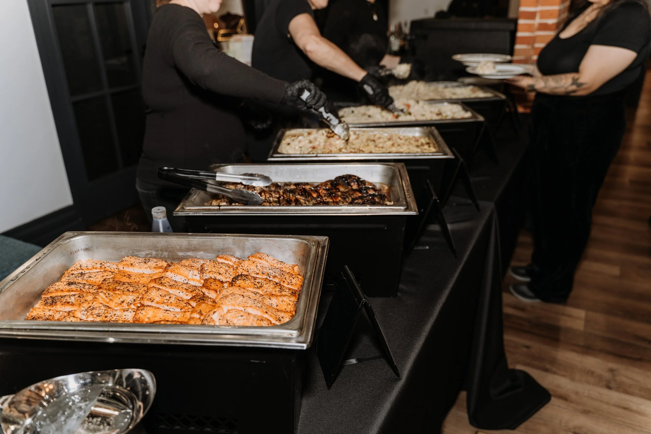 People serving and choosing food from a buffet with various dishes including salmon, grilled chicken, rice, and other cooked foods in metal trays.
