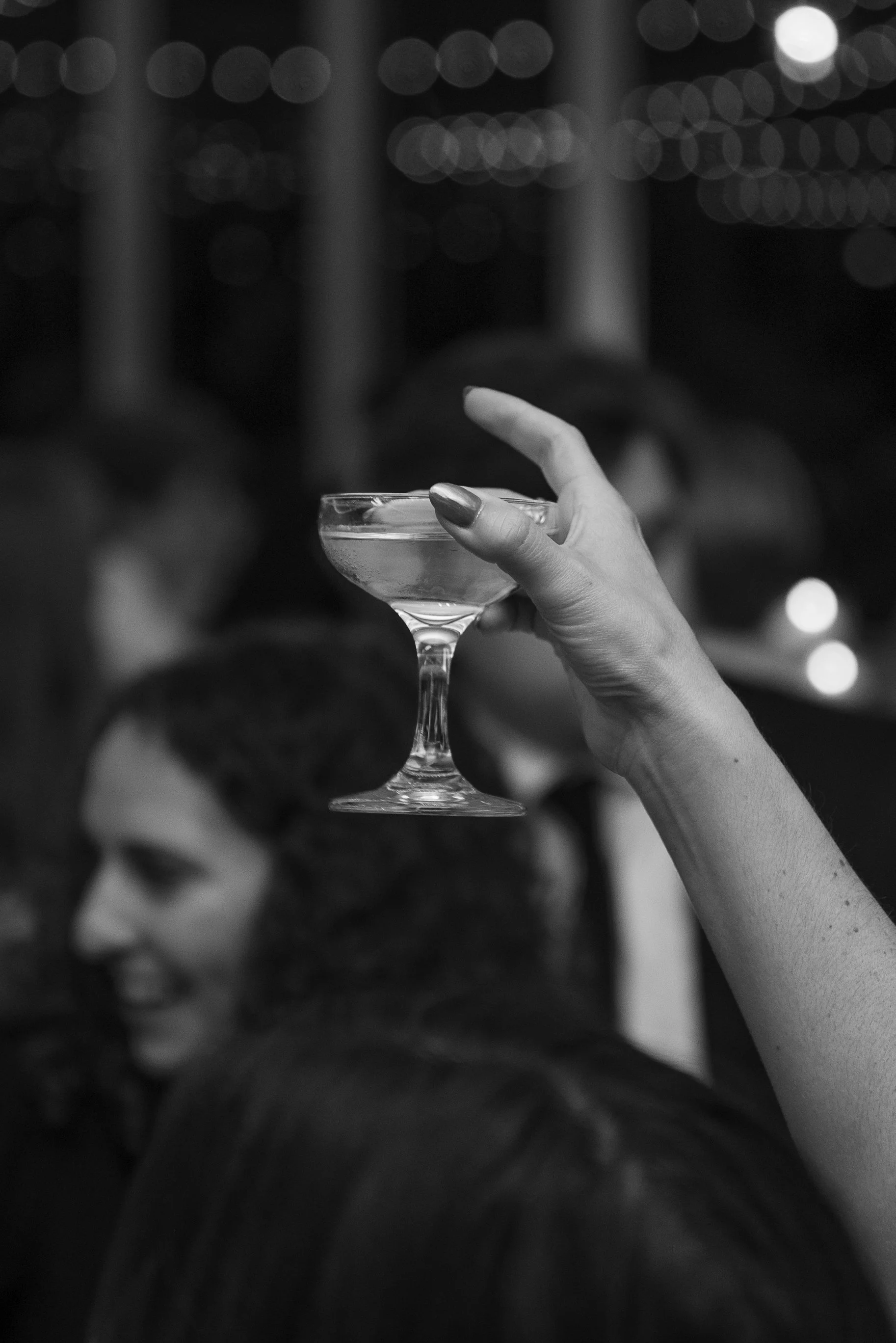A woman's hand holding a cocktail glass in a bar or club setting, with people smiling and blurred in the background.