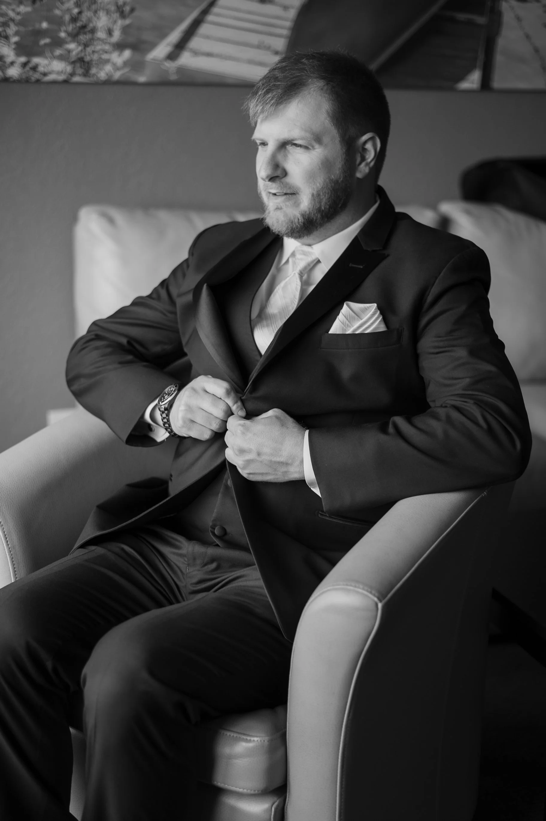 A man in a black suit adjusting his jacket while sitting in a chair, with a white dress shirt, a pocket square, and a watch, in a monochrome photo.