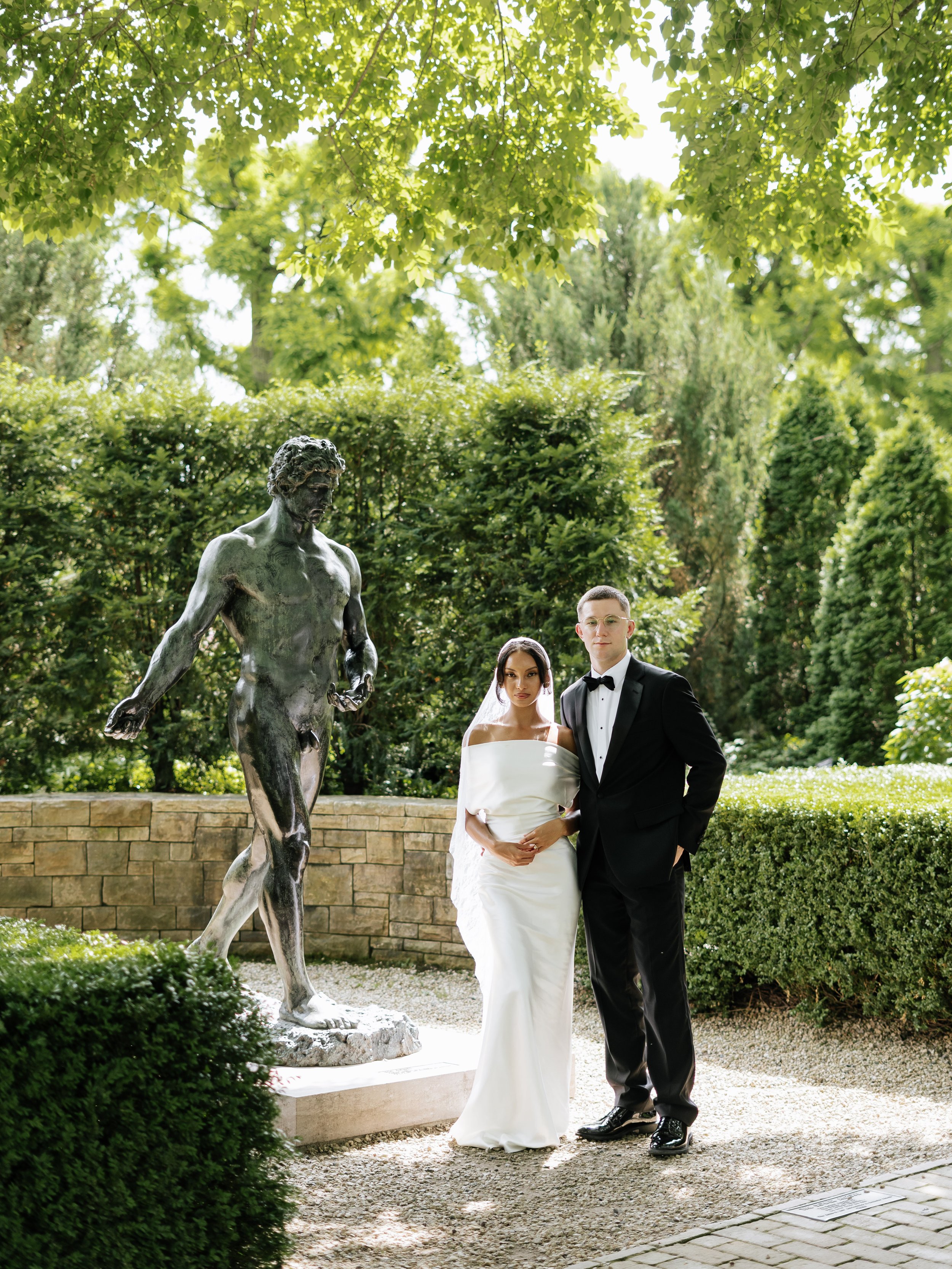 A bride and groom standing outdoors next to a statue of a male figure, with lush green trees and bushes in the background.