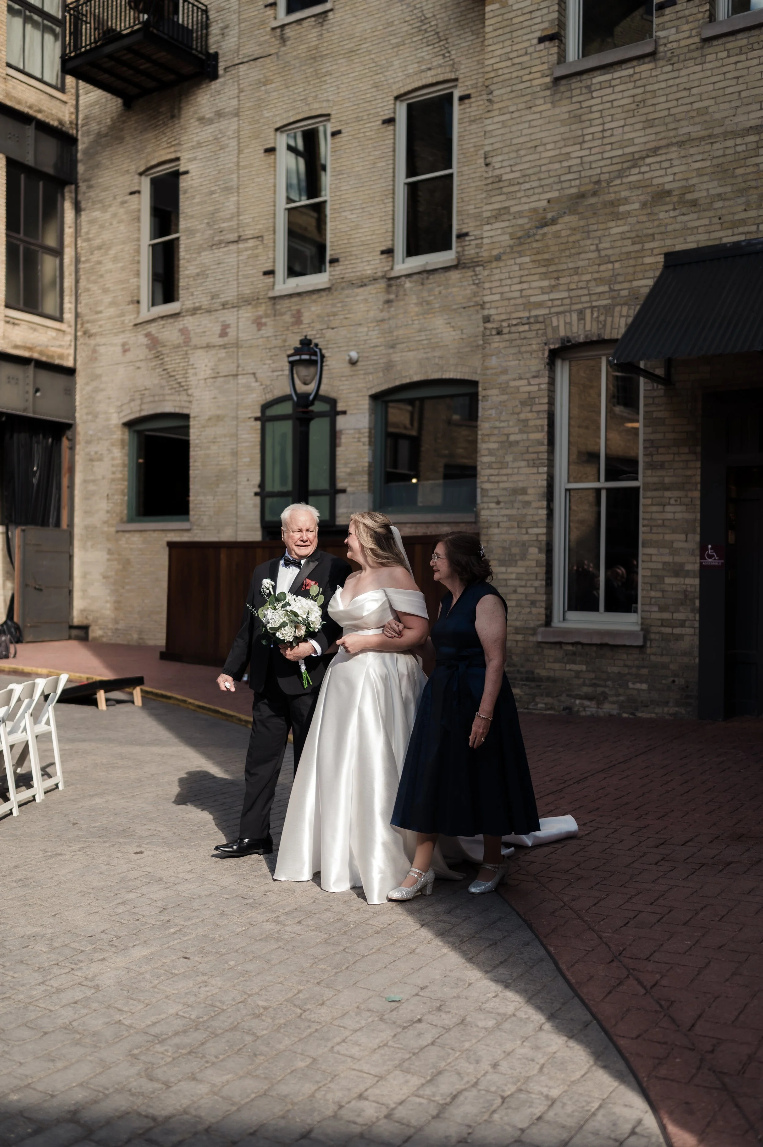 A bride in a white gown walking arm-in-arm with an older man holding a bouquet, accompanied by a woman in a dark dress, outdoors near a brick building during a wedding ceremony or celebration.