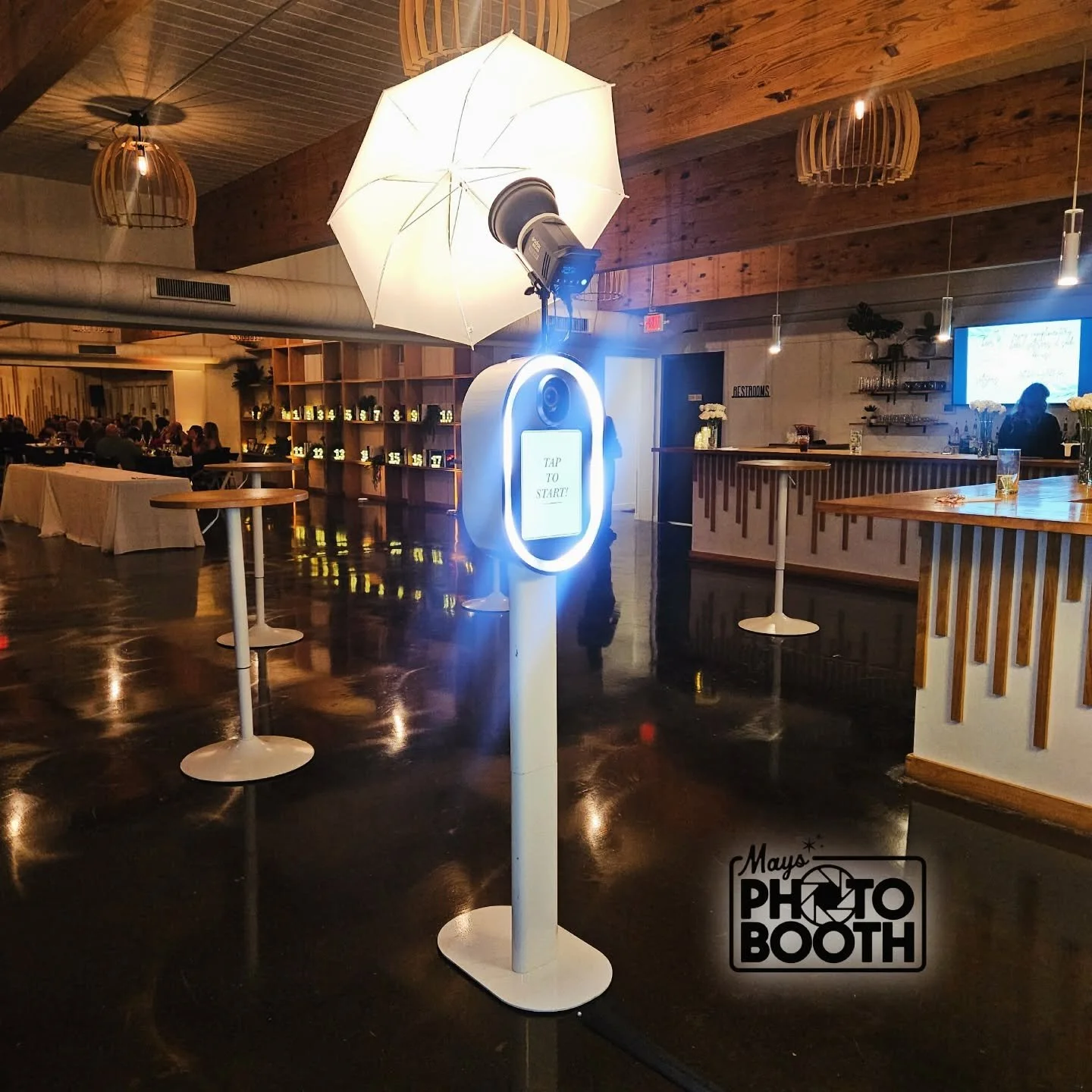 Photo of a photo booth station with a ring light, umbrella, and a sign that says 'Tap to Start' in an event space with tables, a bar area, and a large digital screen in the background.