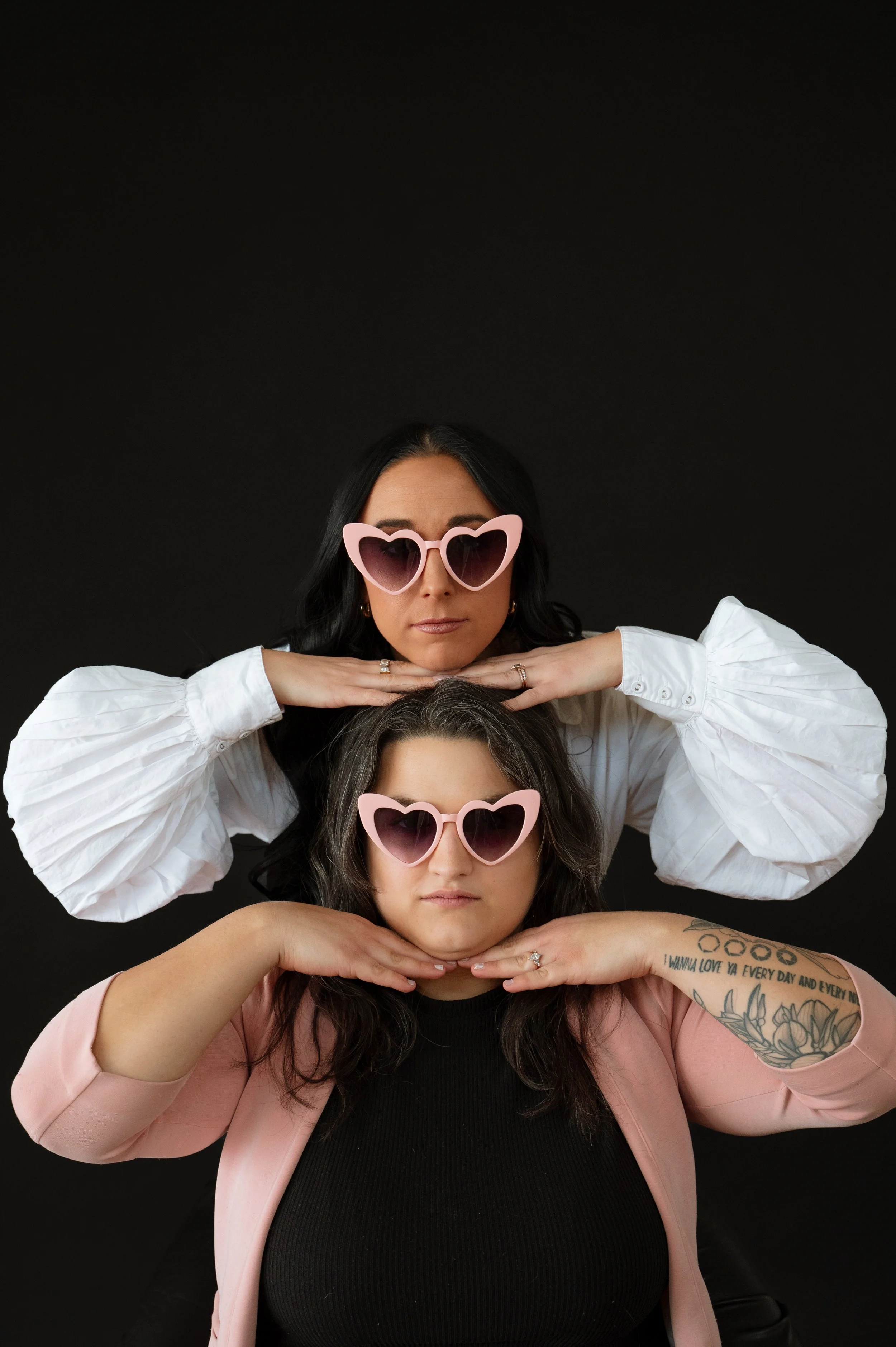 Three women with dark hair and light skin, all wearing pink heart-shaped sunglasses, posing against a black background. They are stacked vertically with their hands under their chins, and the woman at the bottom has tattoos on her right arm.