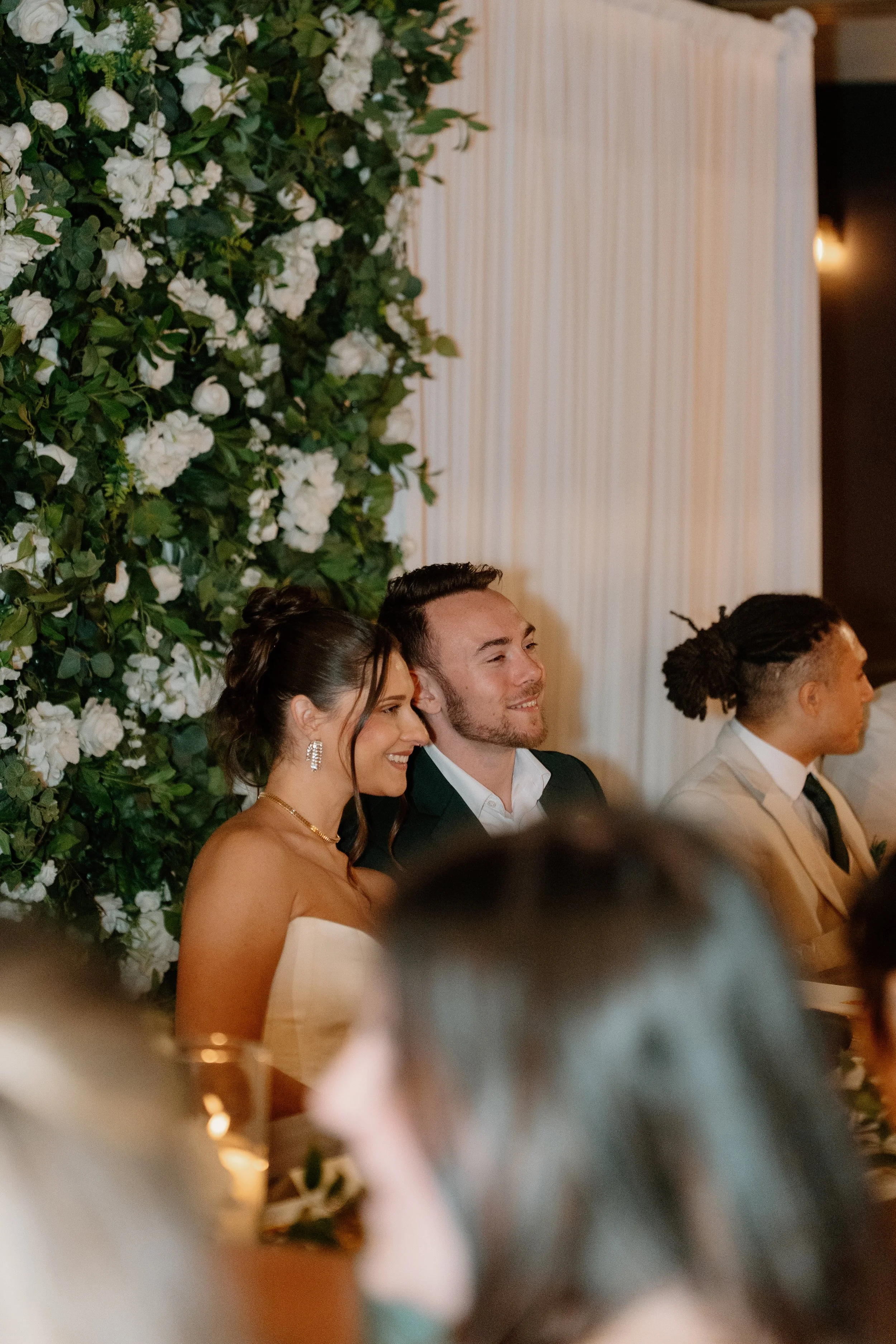 Smiling woman in a strapless dress, next to a man in a suit, at a wedding or formal event, with a floral backdrop and white drapery.