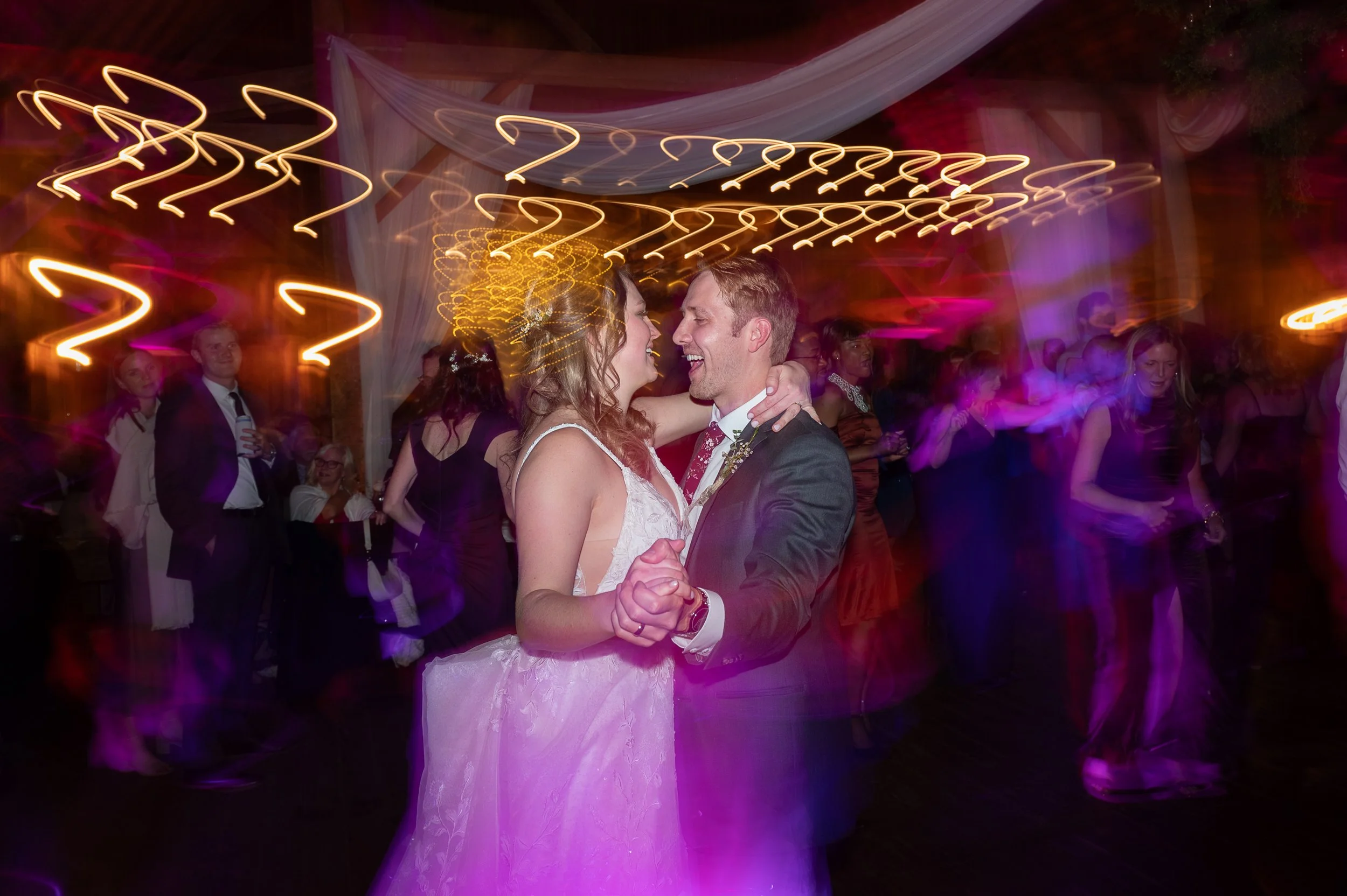 A newlywed couple dancing closely at their wedding reception, with colorful lights and other guests dancing in the background.