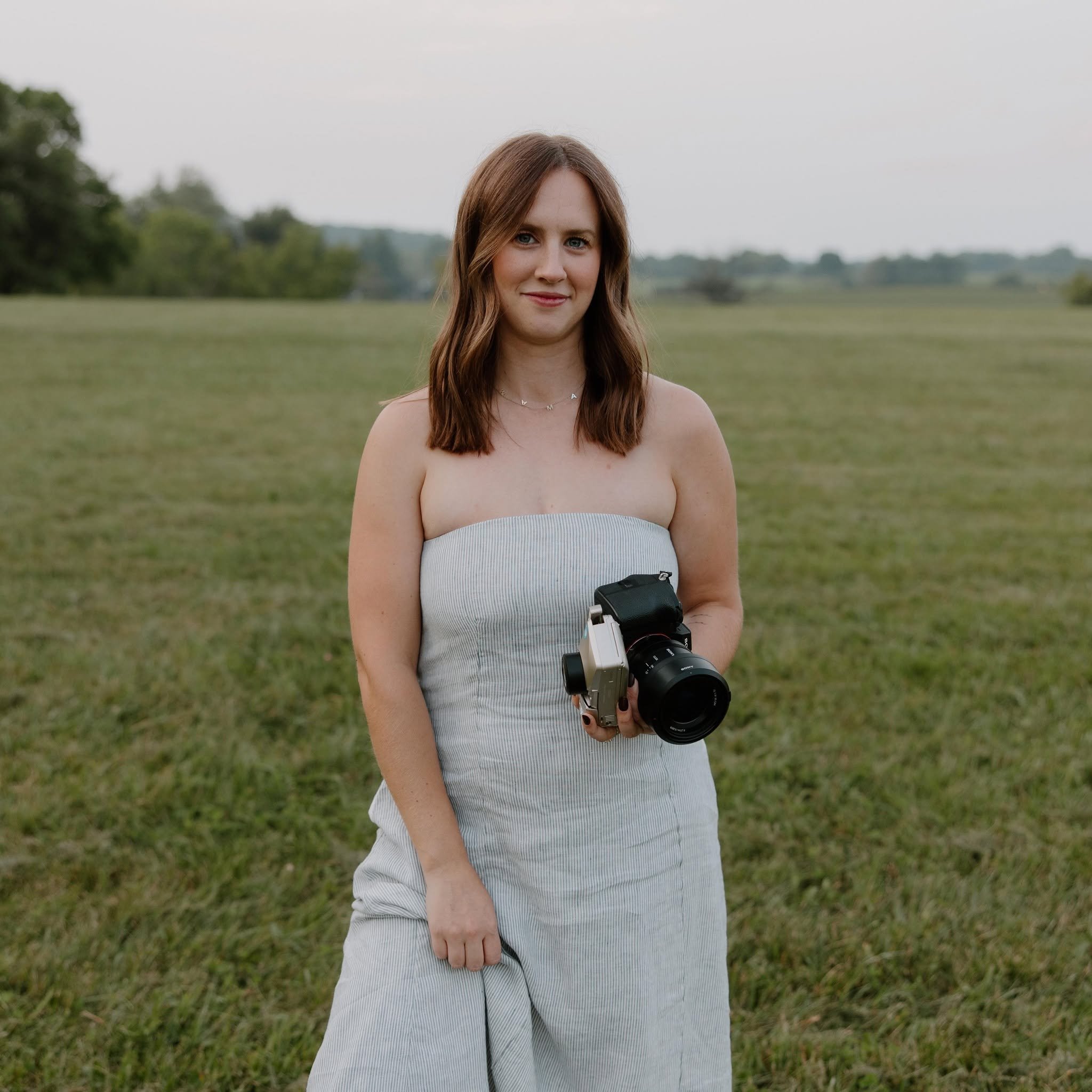A young woman with brown hair and blue eyes standing outdoors in a grassy field on a cloudy day, holding a camera in her right hand.