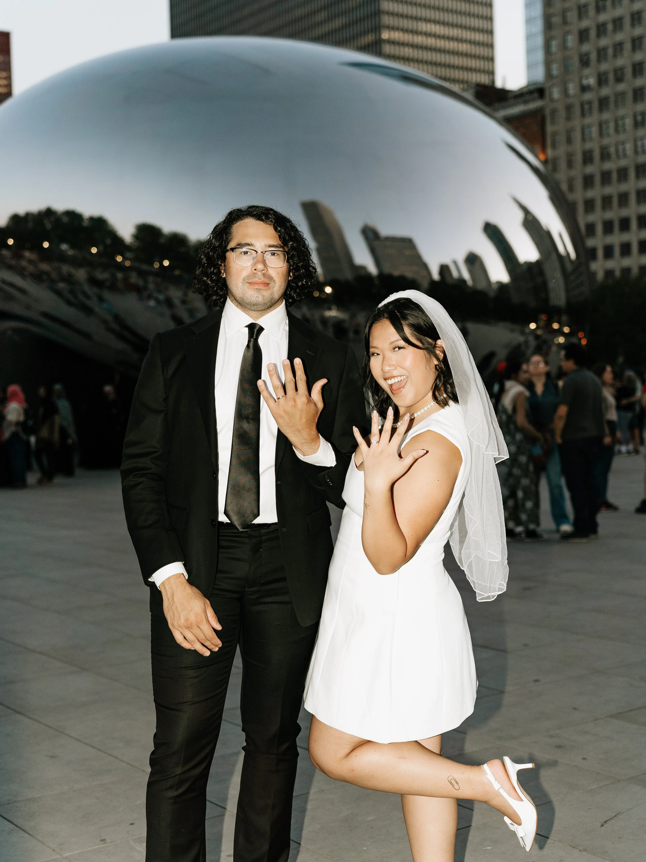 A bride and groom display their wedding rings in front of the Cloud Gate sculpture in Millennium Park, Chicago, during sunset.