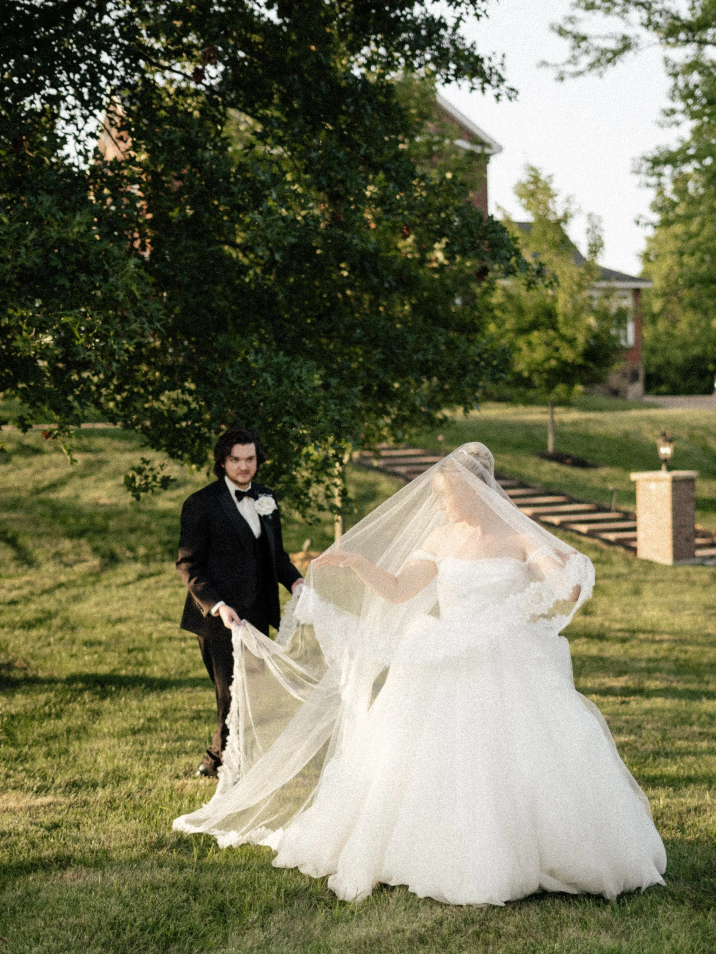 A bride in a white wedding gown and veil is being assisted by a groom in a black tuxedo with a bow tie and white boutonniere outside on a grassy area near trees and houses.