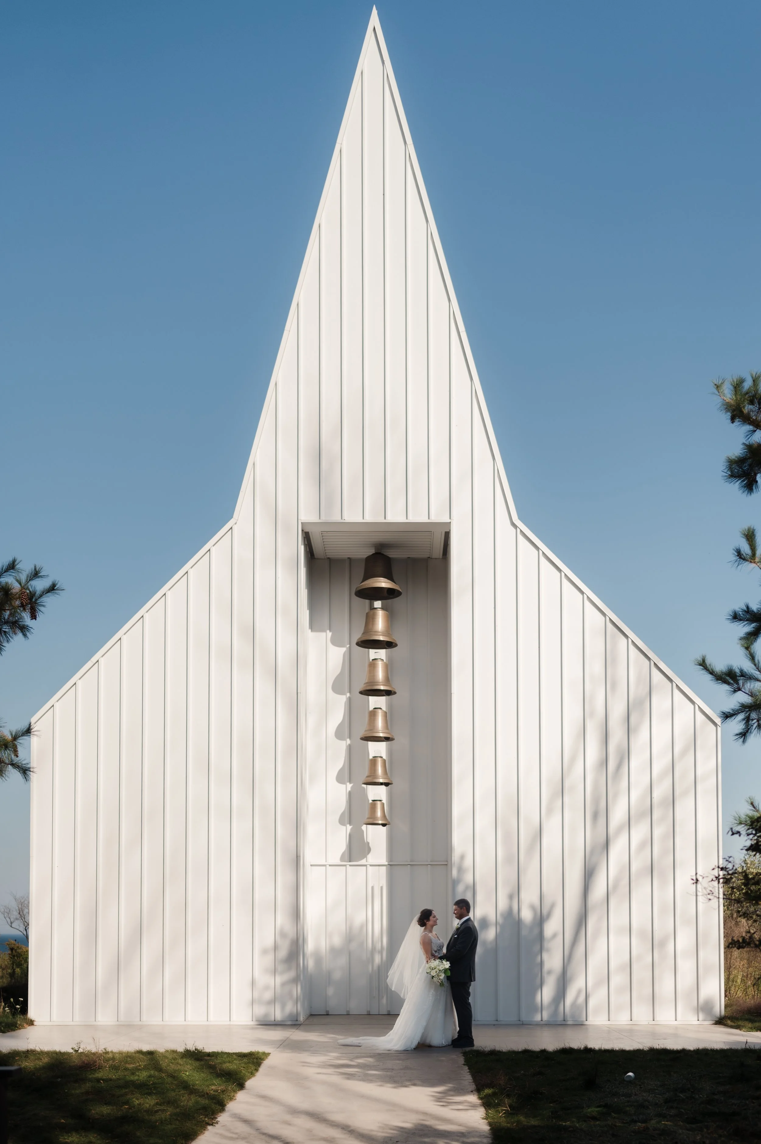 A bride and groom standing and facing each other in front of a modern, white church with a tall, pointed roof and hanging bells, outdoors on a sunny day.