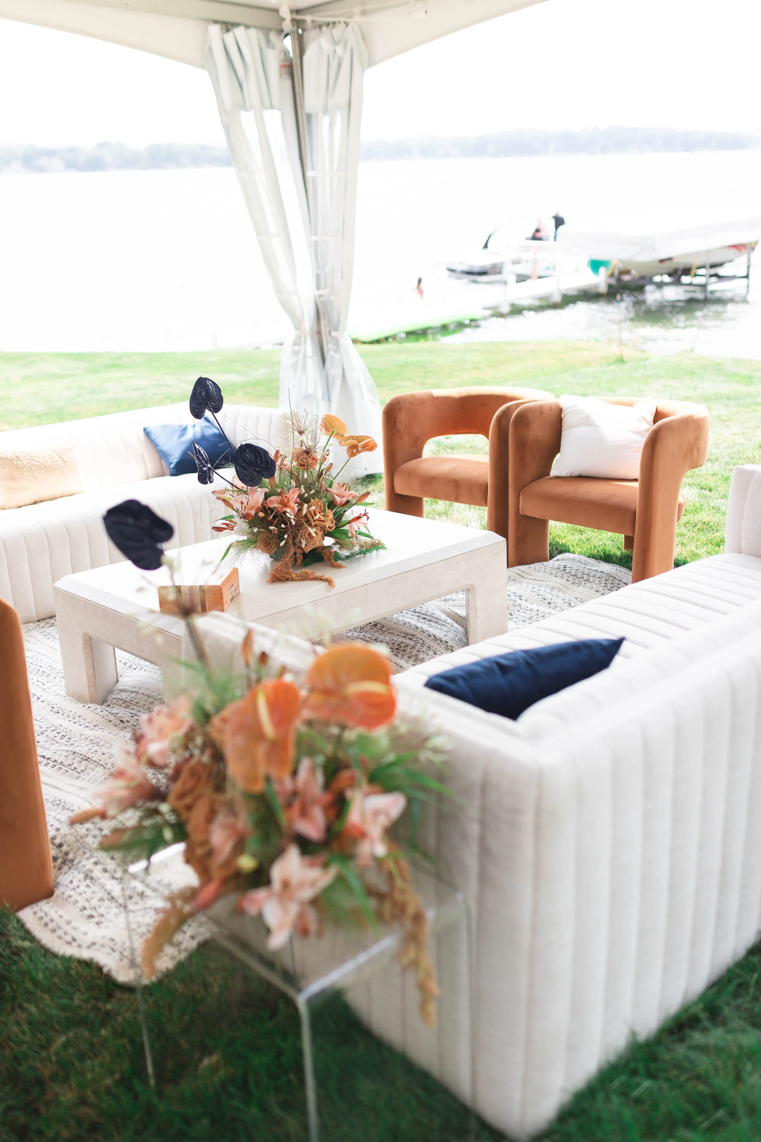 A cozy outdoor seating area on a grass lawn with a view of a lake and boats in the background, featuring beige sofas with dark pillows, wooden chairs, a white coffee table with a floral arrangement, and a white canopy.