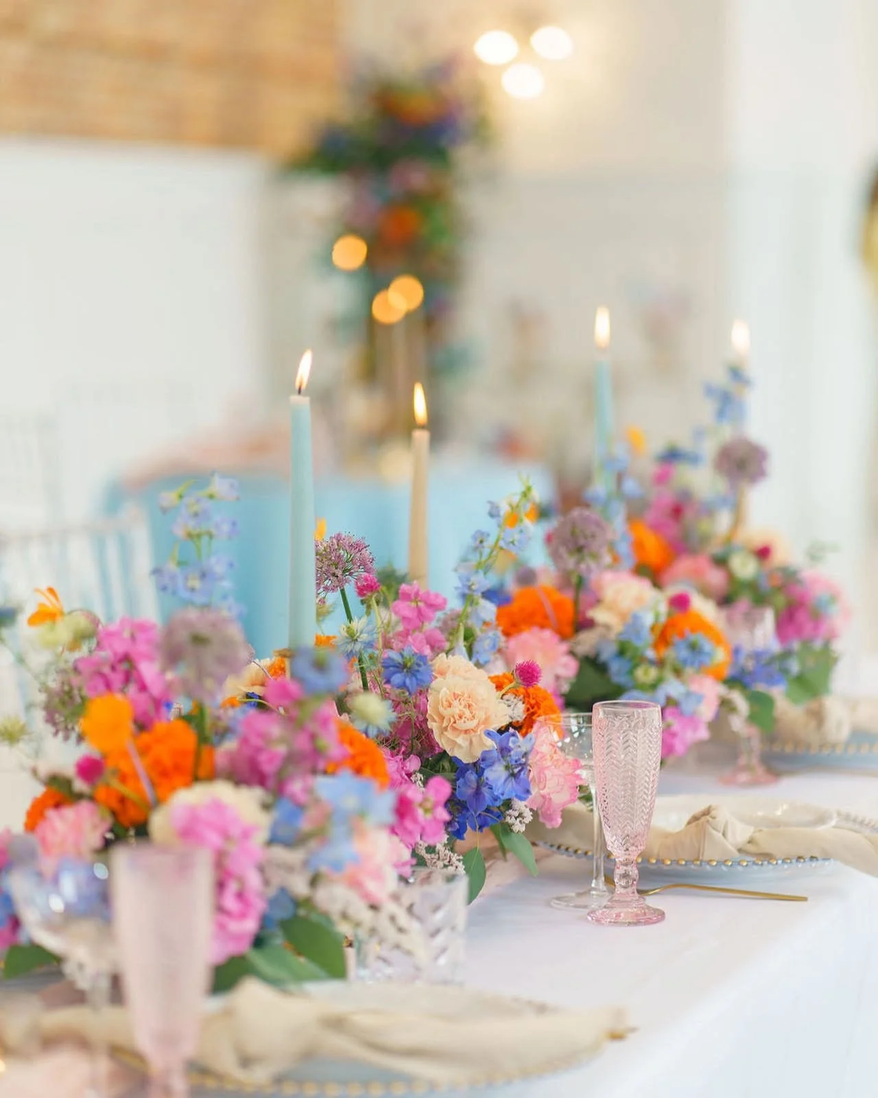 A table decorated with colorful flowers including pink, orange, purple, and white blooms, lit candles, and elegant pink glassware, set for a festive event or celebration.