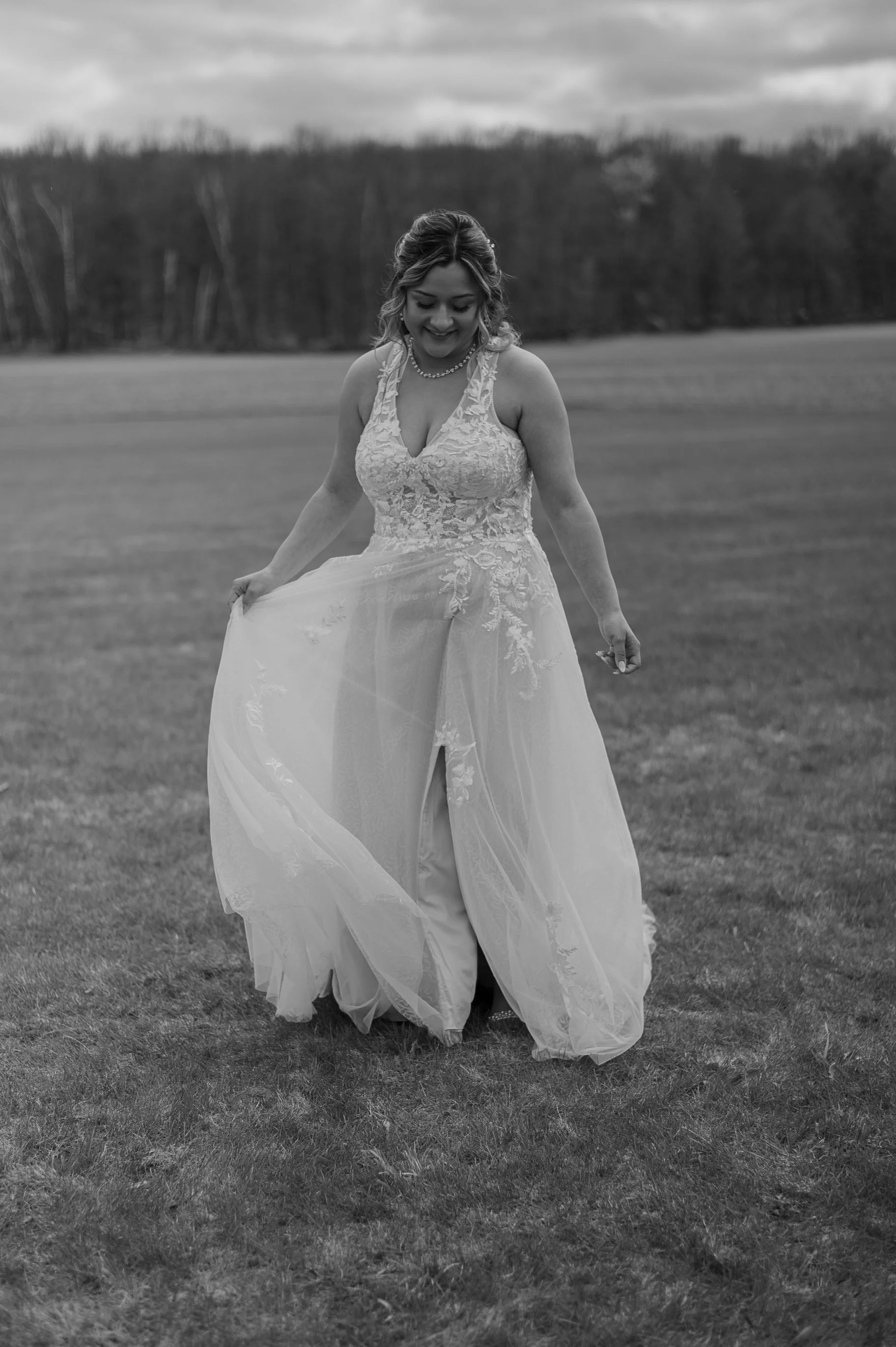 A woman in a wedding dress standing on a grassy field, holding the skirt of her dress, with trees and a cloudy sky in the background.