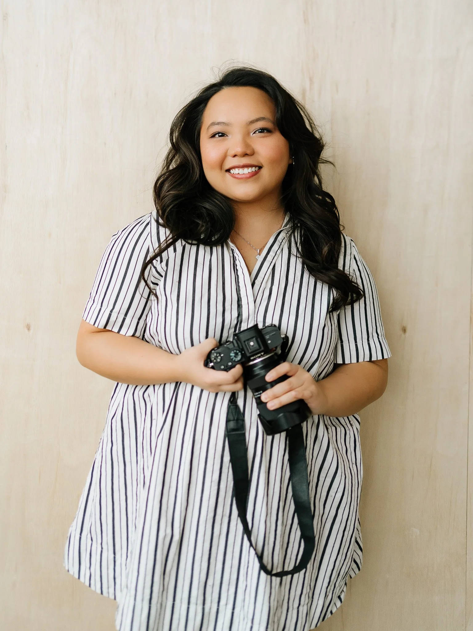A young woman sitting on a cream-colored sofa, holding a camera and smiling at the camera, in a cozy room with brick walls and sheer curtains.
