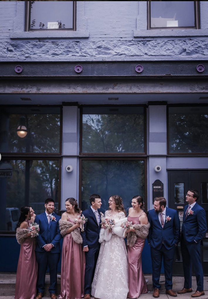 A wedding party stands outside a building with large windows, including the bride in a white lace gown holding a bouquet, surrounded by bridesmaids in pink dresses with fur stoles and groomsmen in blue suits with pink boutonnieres, all smiling and looking at each other.